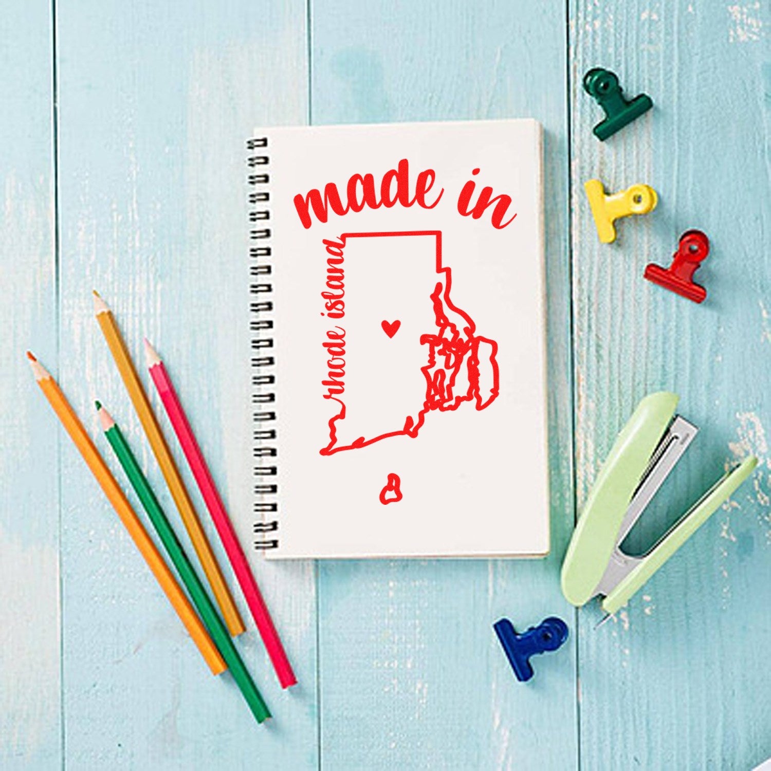 Self-Inking Handmade with Love in Rhode Island Stamp on a notebook with colorful pencils and clips on a blue wooden table.