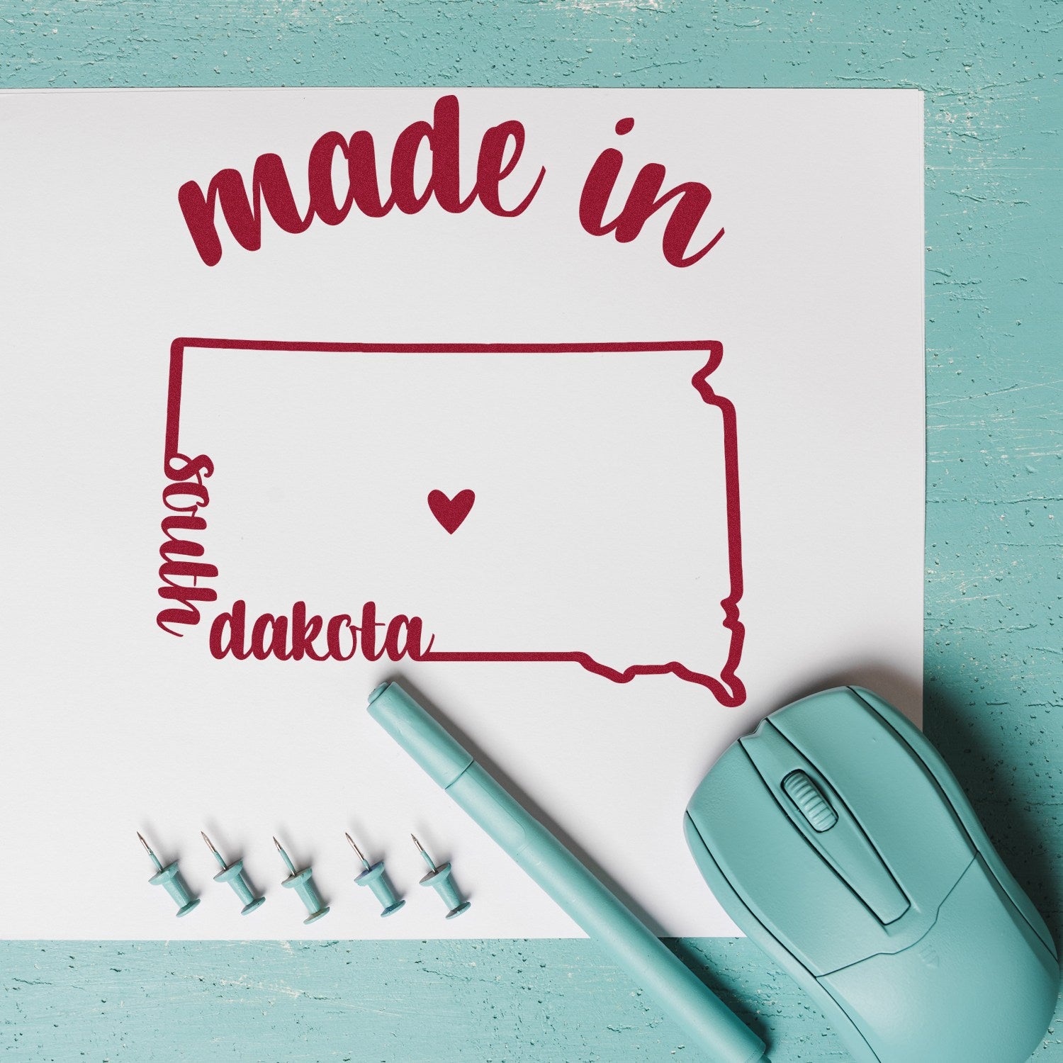 Slim Pre-Inked Stamp South Dakota Made in Stamp on paper with a heart design, surrounded by a teal pen, mouse, and push pins on a turquoise surface.