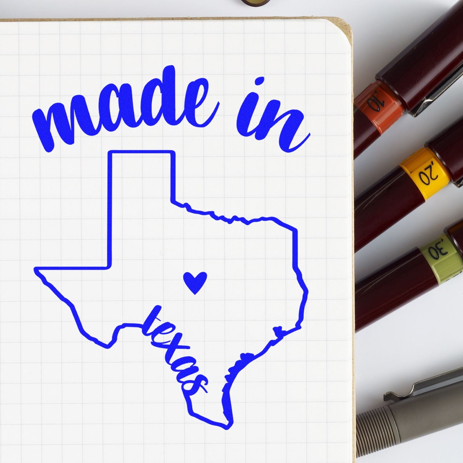 Slim Pre-Inked Stamp Texas Made in Stamp featuring a blue Texas outline with made in Texas text and heart, shown on a notepad with pens nearby. Perfect for adding a Texan touch.