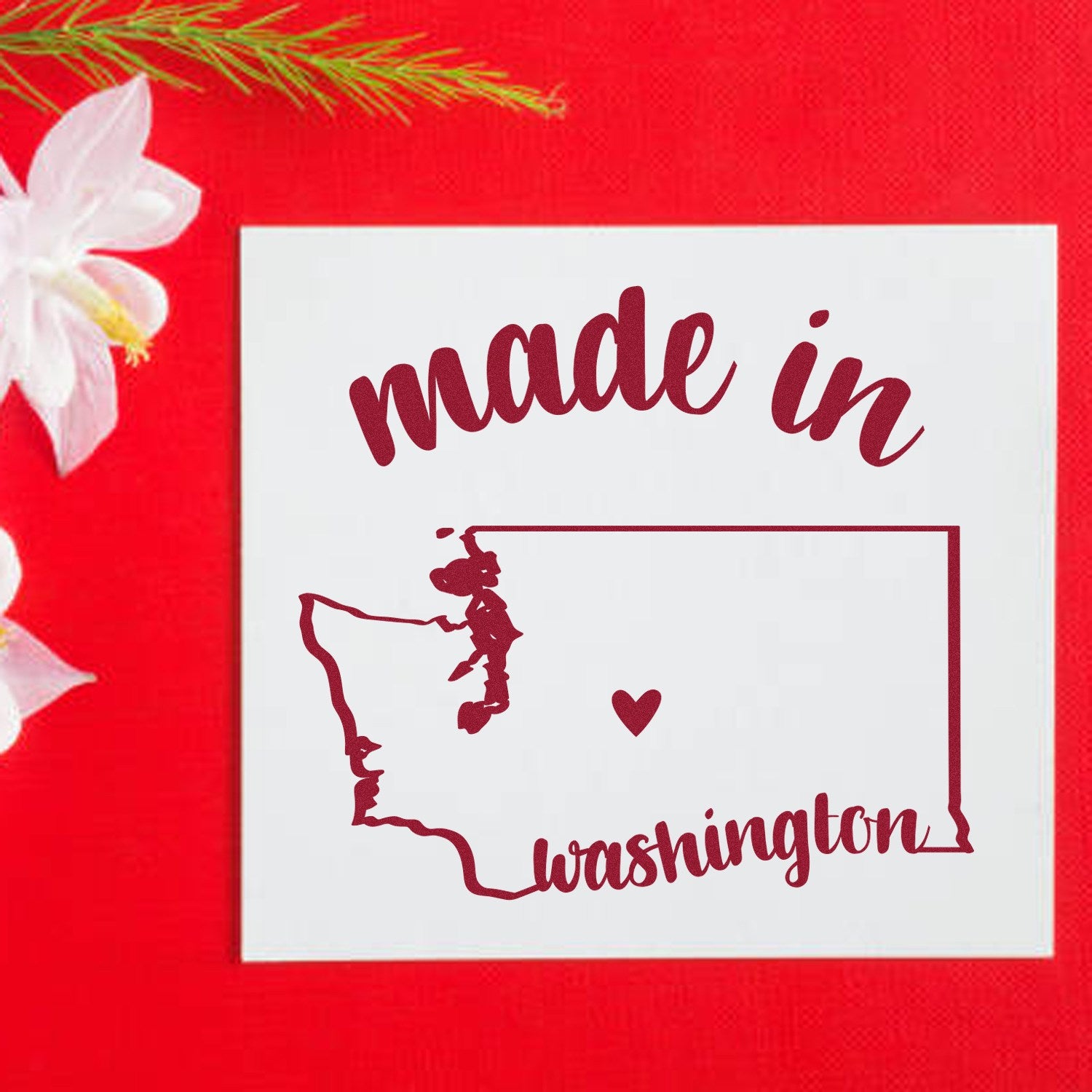 Made in Washington Stamp Pre-Inked: Red stamp with 'made in Washington' text and state outline, featuring a heart. Perfect for crafts and branding. Set against a red background with flowers.