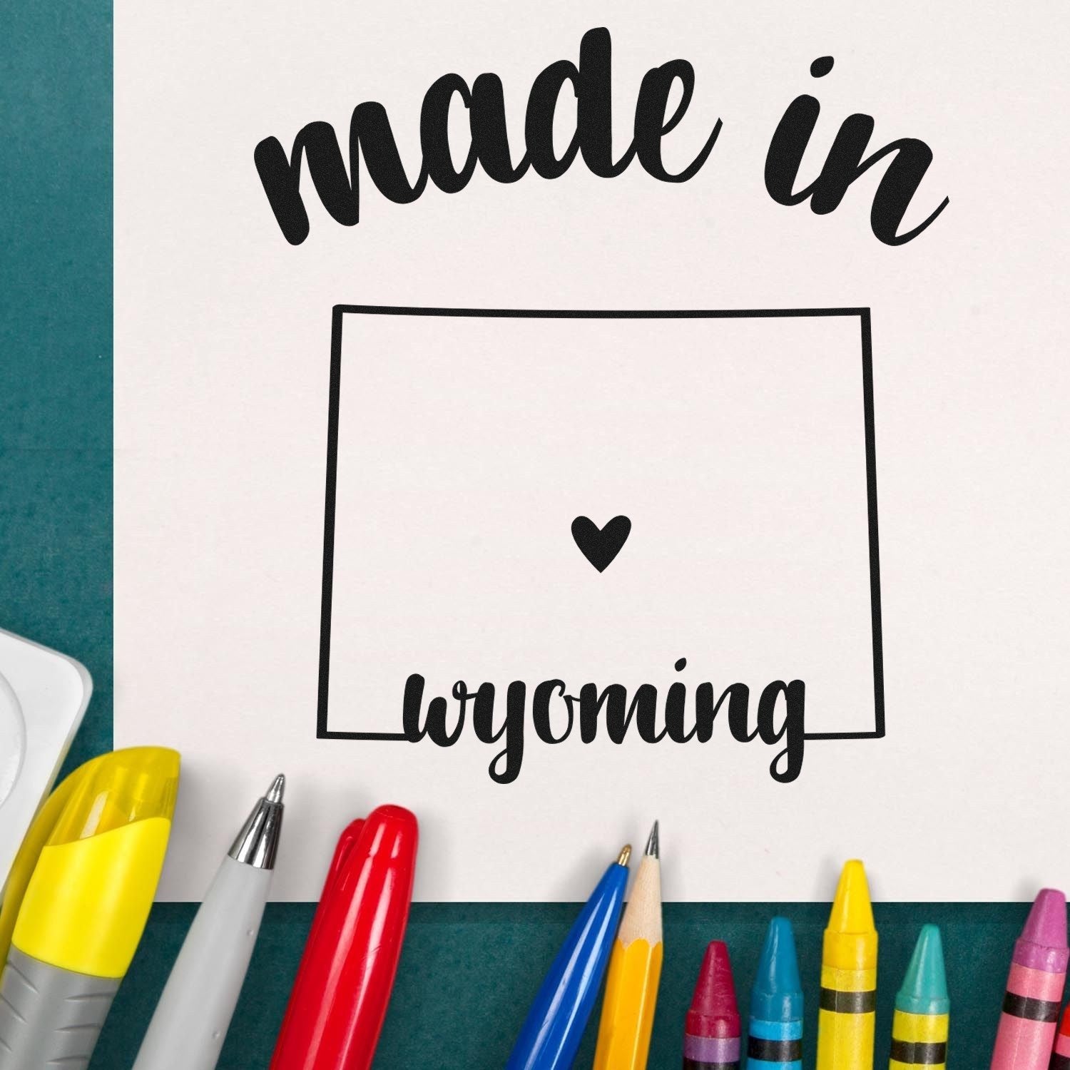 Made with Love in Wyoming Rubber Stamp featuring bold text and a heart inside a Wyoming outline, surrounded by colorful pens and crayons on a green background.