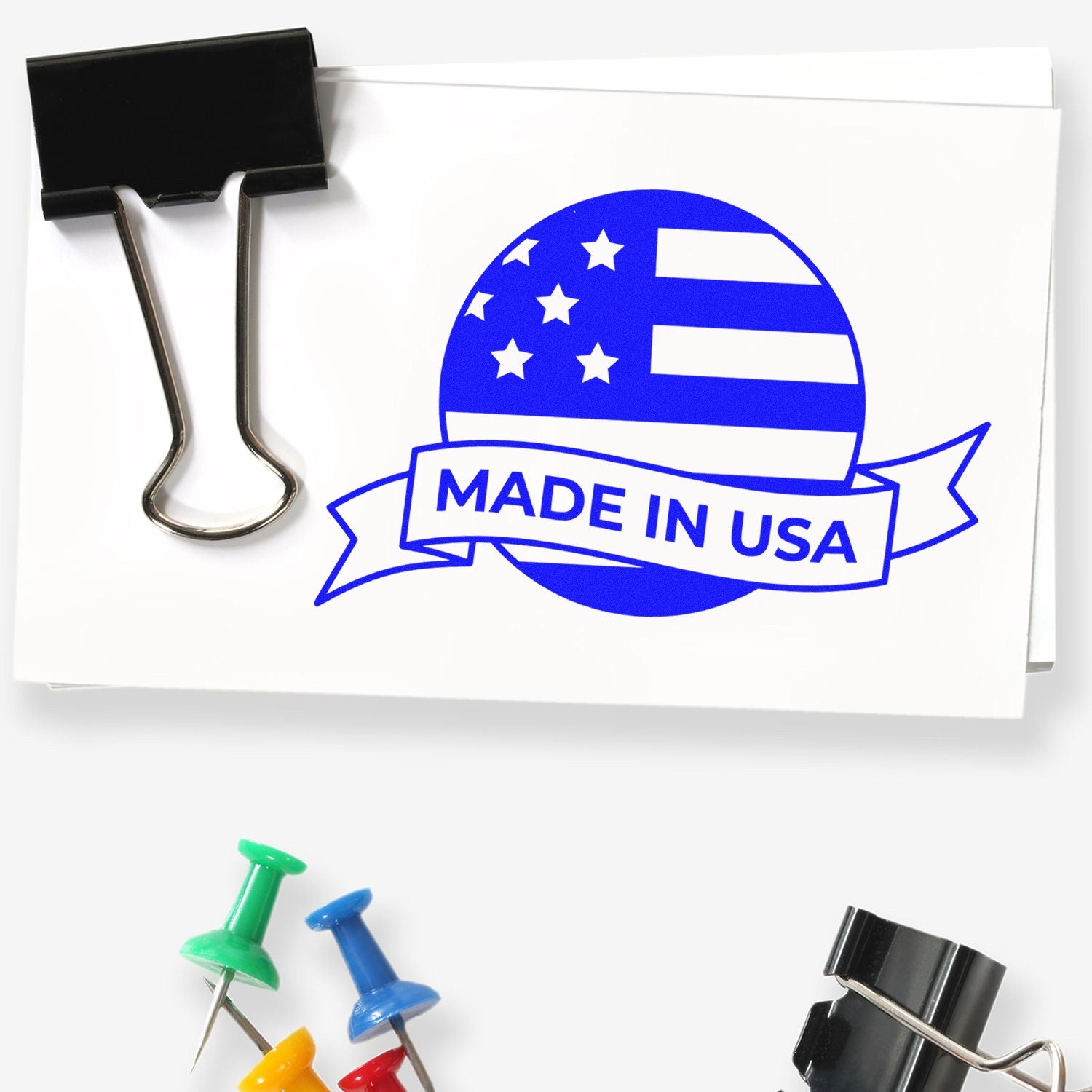 Image of the Slim Pre-Inked Patriot Seal Of Quality Stamp, featuring a blue Made in USA design with stars and stripes, next to colorful push pins and a black binder clip.