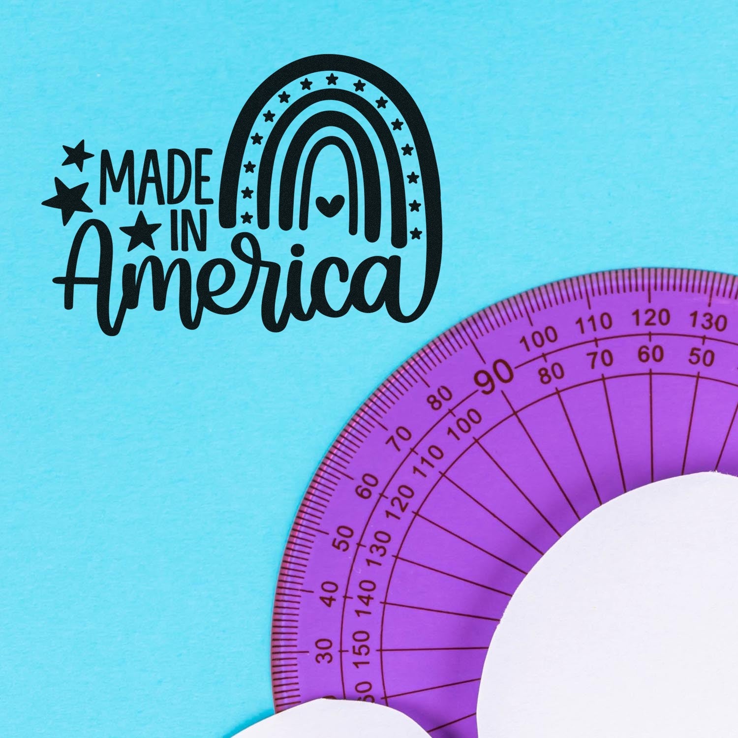 Image of the Slim Pre-Inked American Pride Rainbow Stamp featuring a black rainbow design with stars and Made in America text on a light blue background, next to a purple protractor.