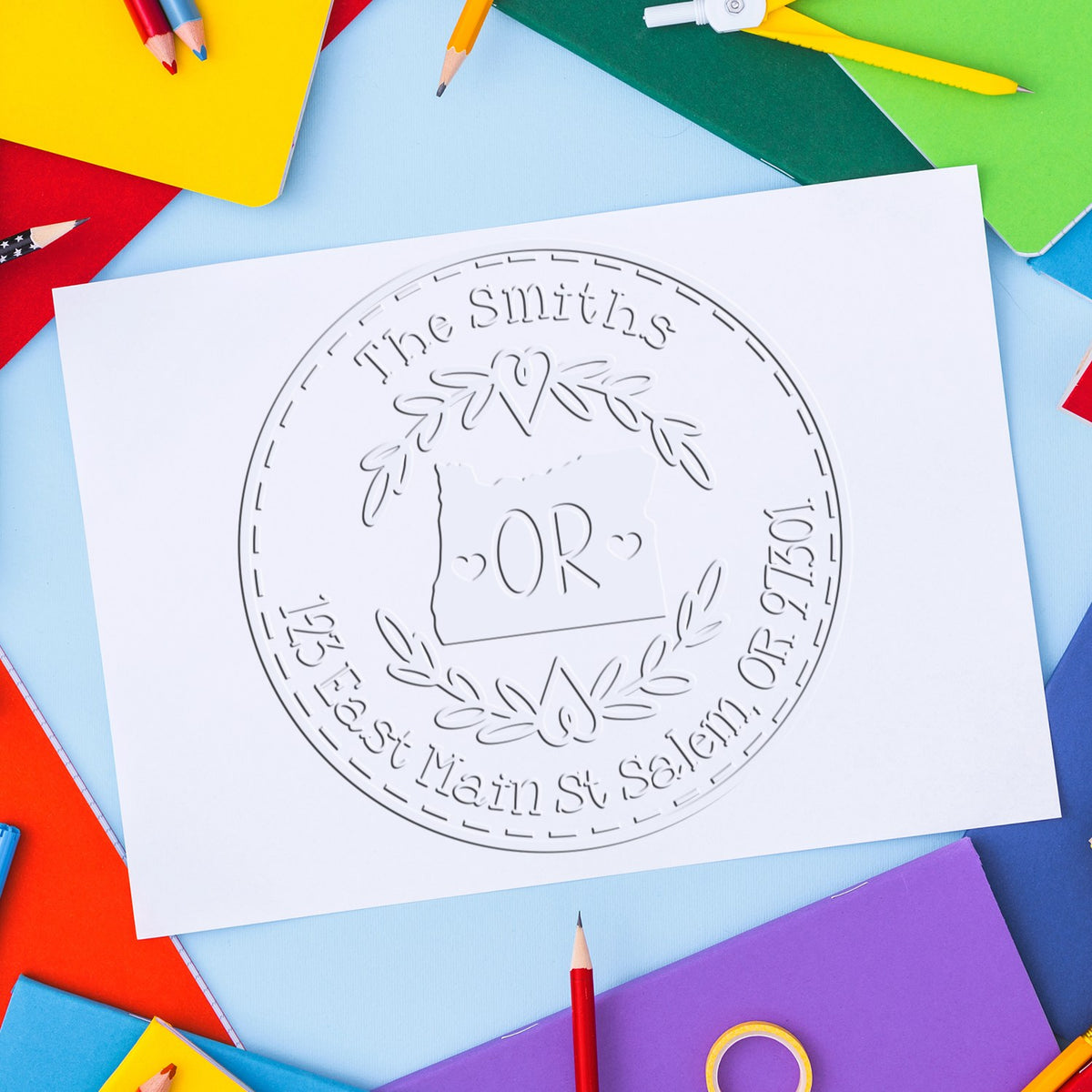 White embossed custom seal stamp on paper, surrounded by colorful notebooks and pencils on blue background