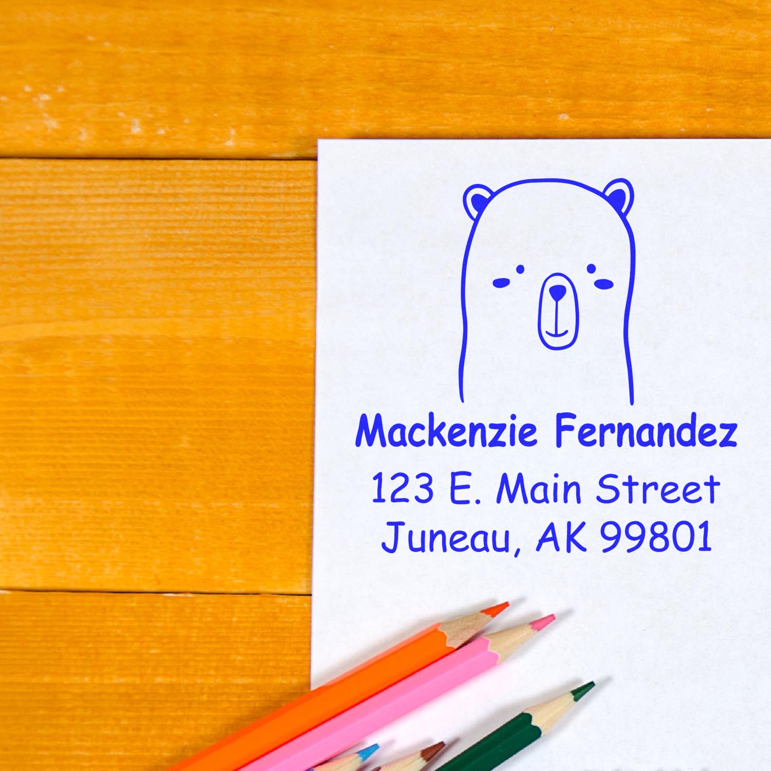 Wood handle adorable bear cartoon animal made-to-order mail stamp on paper with personalized address stamp reading "Mackenzie Fernandez, 123 E. Main Street, Juneau, AK 99801." Colored pencils are nearby on a wooden surface.
