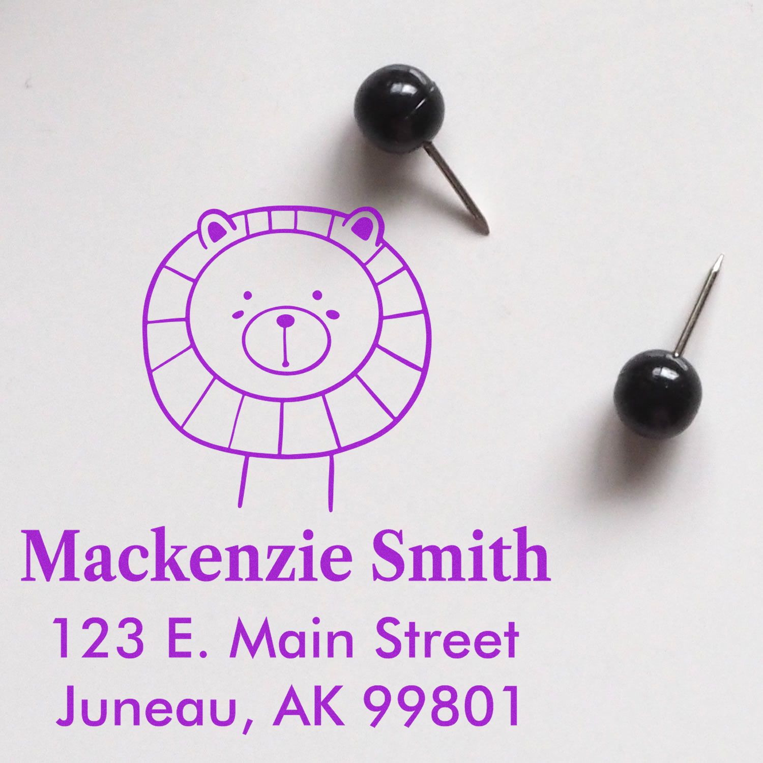 Wood Handle Lovable Lion's Lair Cartoon Animal Made To Order Return Address Stamp featuring a cute lion design. Customizable Address Stamp with the name "Mackenzie Smith" and address "123 E. Main Street, Juneau, AK 99801" in purple text, next to two black push pins.