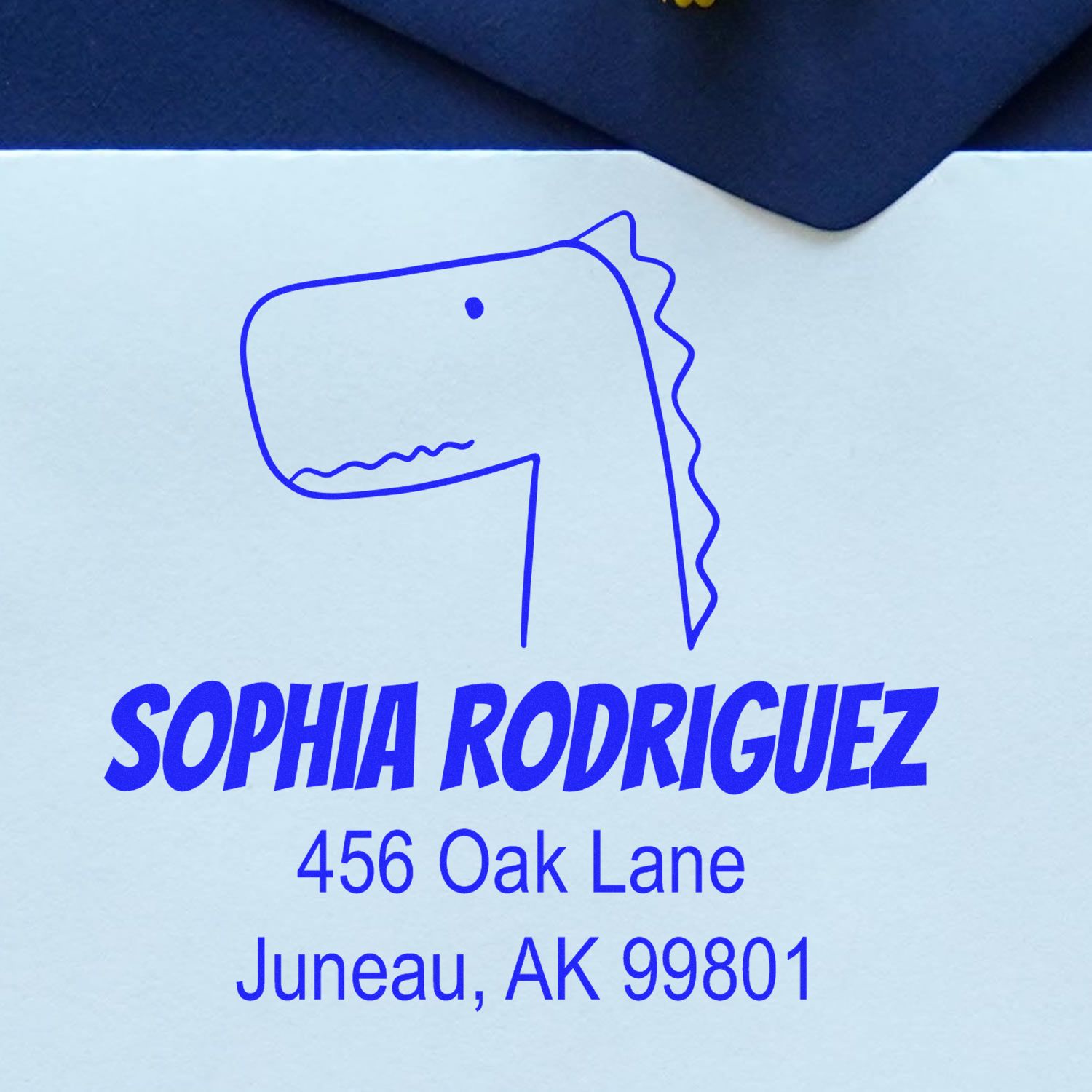 Wood handle Mirthful Dino cartoon animal made-to-order address stamp on an envelope. The customized new address stamp features a playful dinosaur design with the name "Sophia Rodriguez" and address "456 Oak Lane, Juneau, AK 99801" in blue ink.