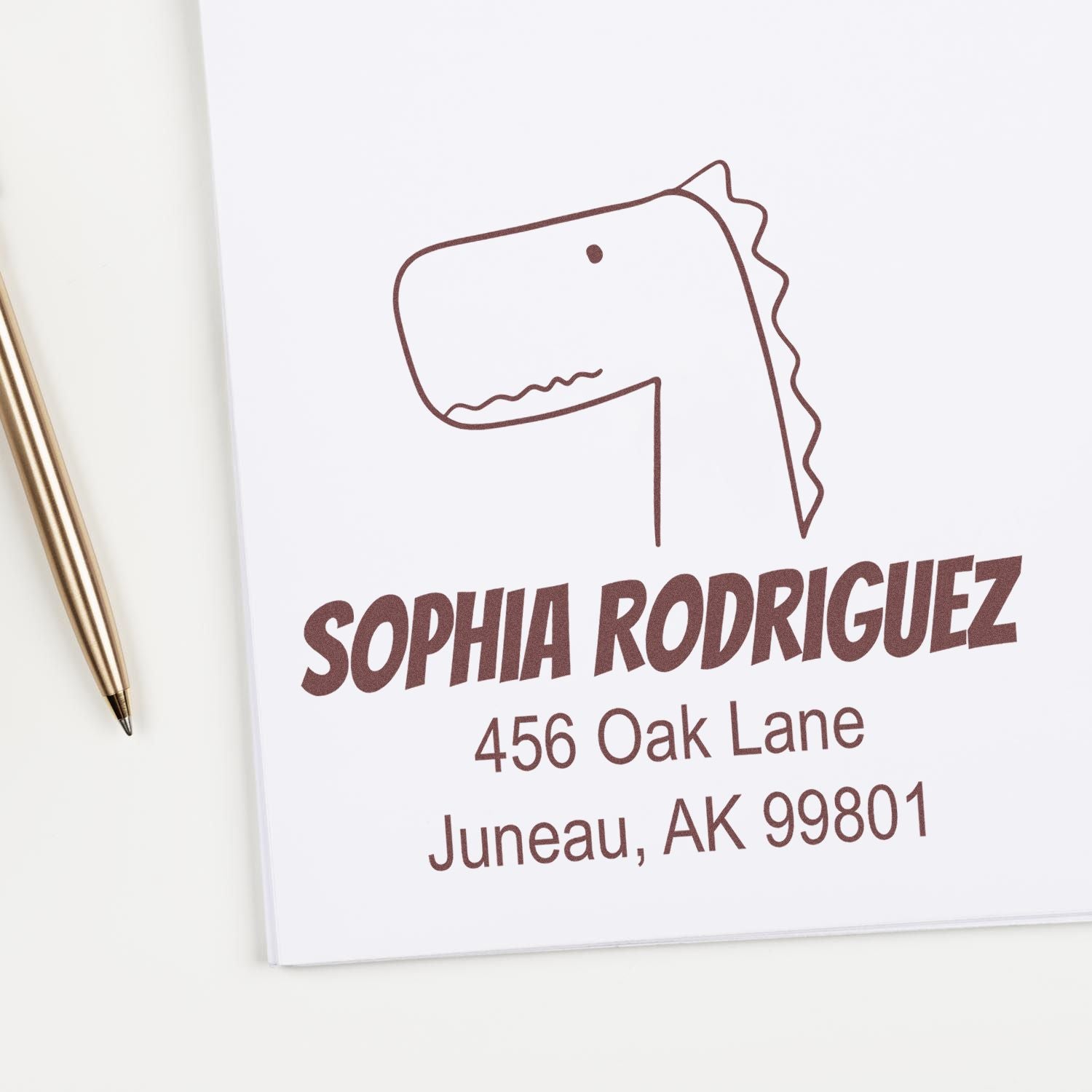 Portable Pre-Inked Mirthful Dino Custom Designed Mail Stamper in use, featuring a customizable address stamp with a dinosaur illustration. The stamp displays the name "Sophia Rodriguez" and an address in Juneau, AK. A pen is placed nearby.