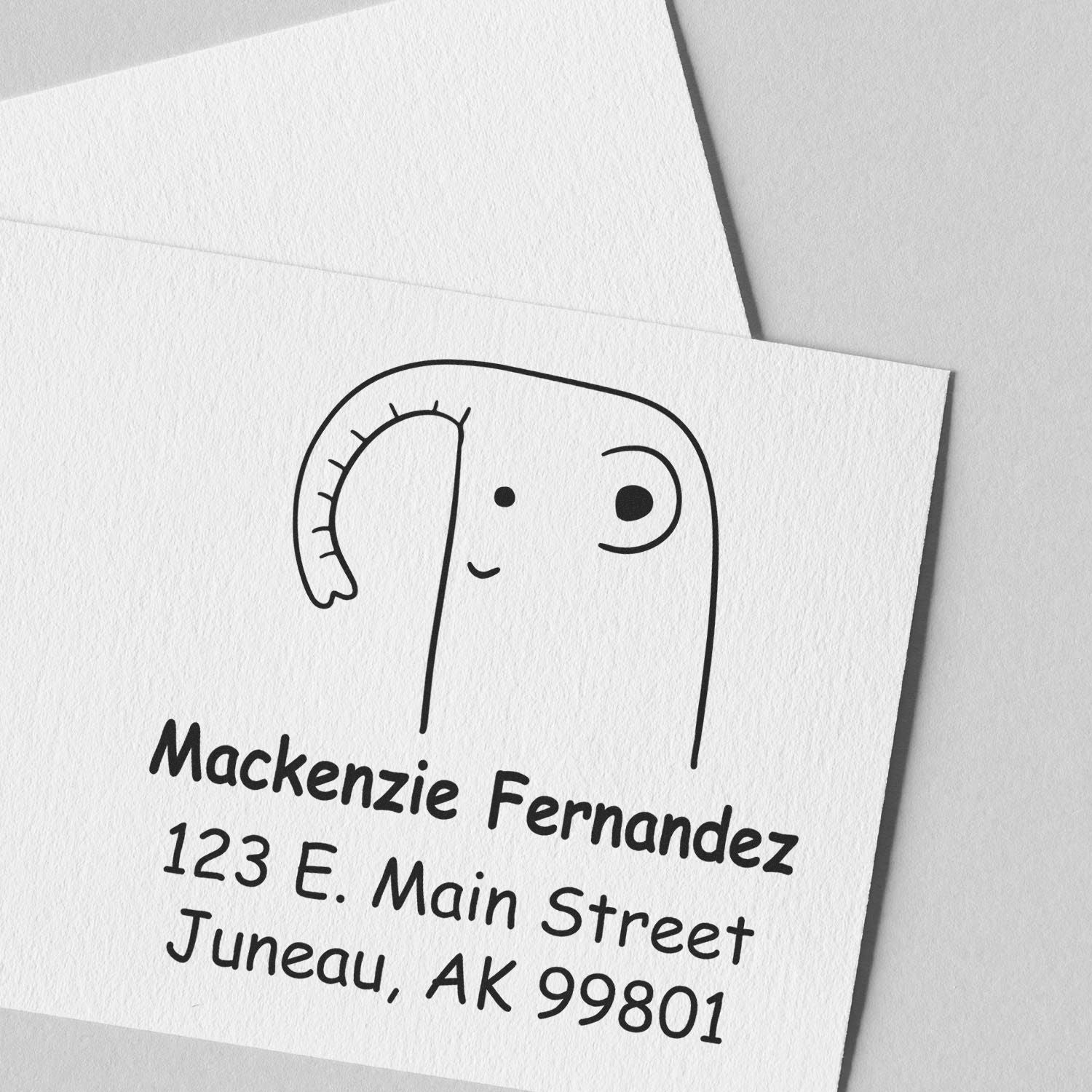 Endearing elephant cartoon animal with a wood handle on a custom-made mailing stamp. The address stamper features "Mackenzie Fernandez, 123 E. Main Street, Juneau, AK 99801" in black ink on white paper.