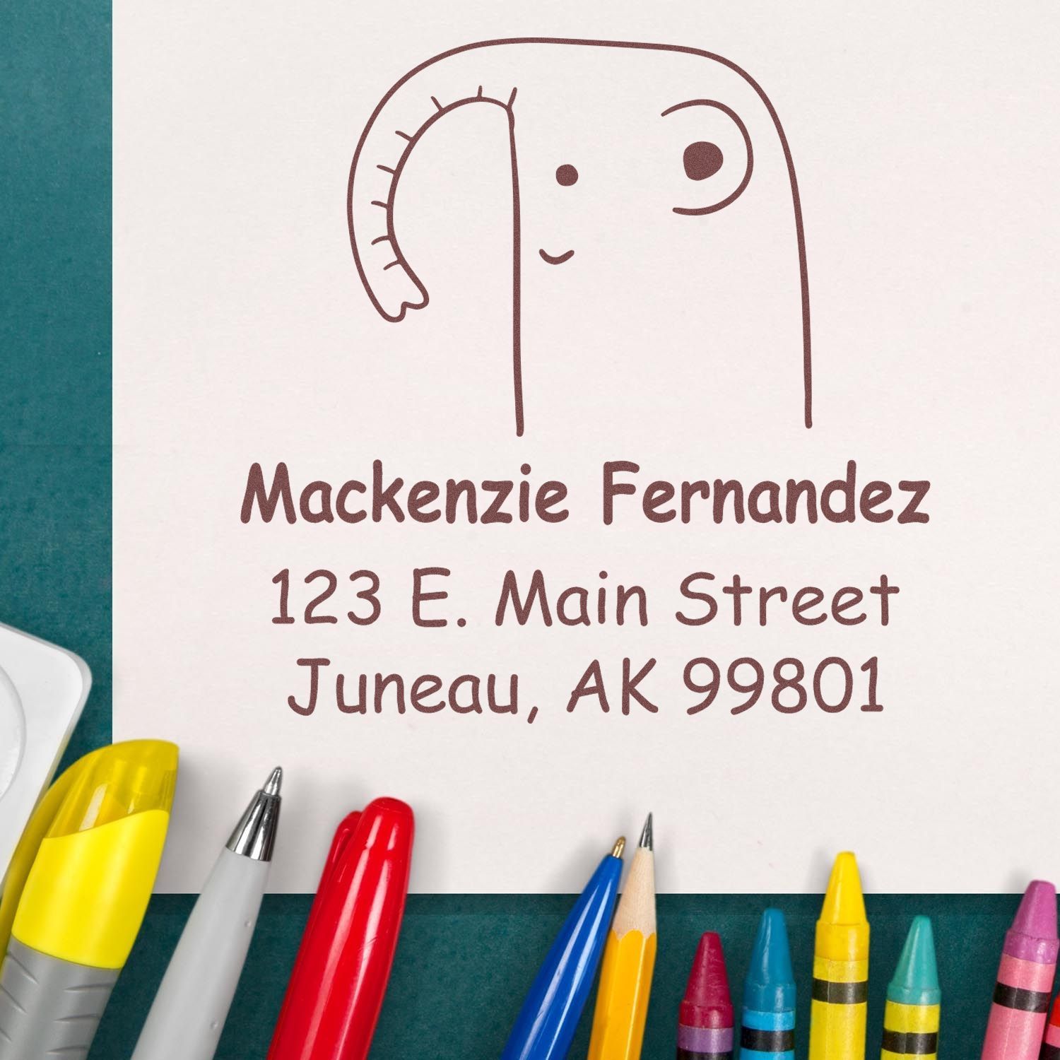 Endearing self-inking cartoon animal elephant custom made address stamp on paper with "Mackenzie Fernandez, 123 E. Main Street, Juneau, AK 99801." Surrounded by colorful pens and crayons, showcasing a custom return mail stamp design.
