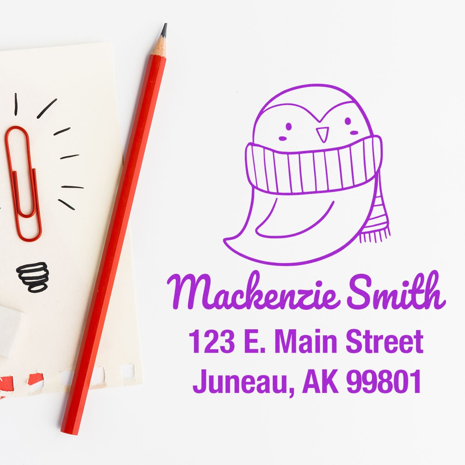 Self-Inking Cartoon Animal Pretty Penguin Custom Made Address Stamper featuring a cute penguin in a scarf. The Customized Mailing Stamp displays the name Mackenzie Smith and address in purple ink, next to a red pencil and paper with a red paperclip.