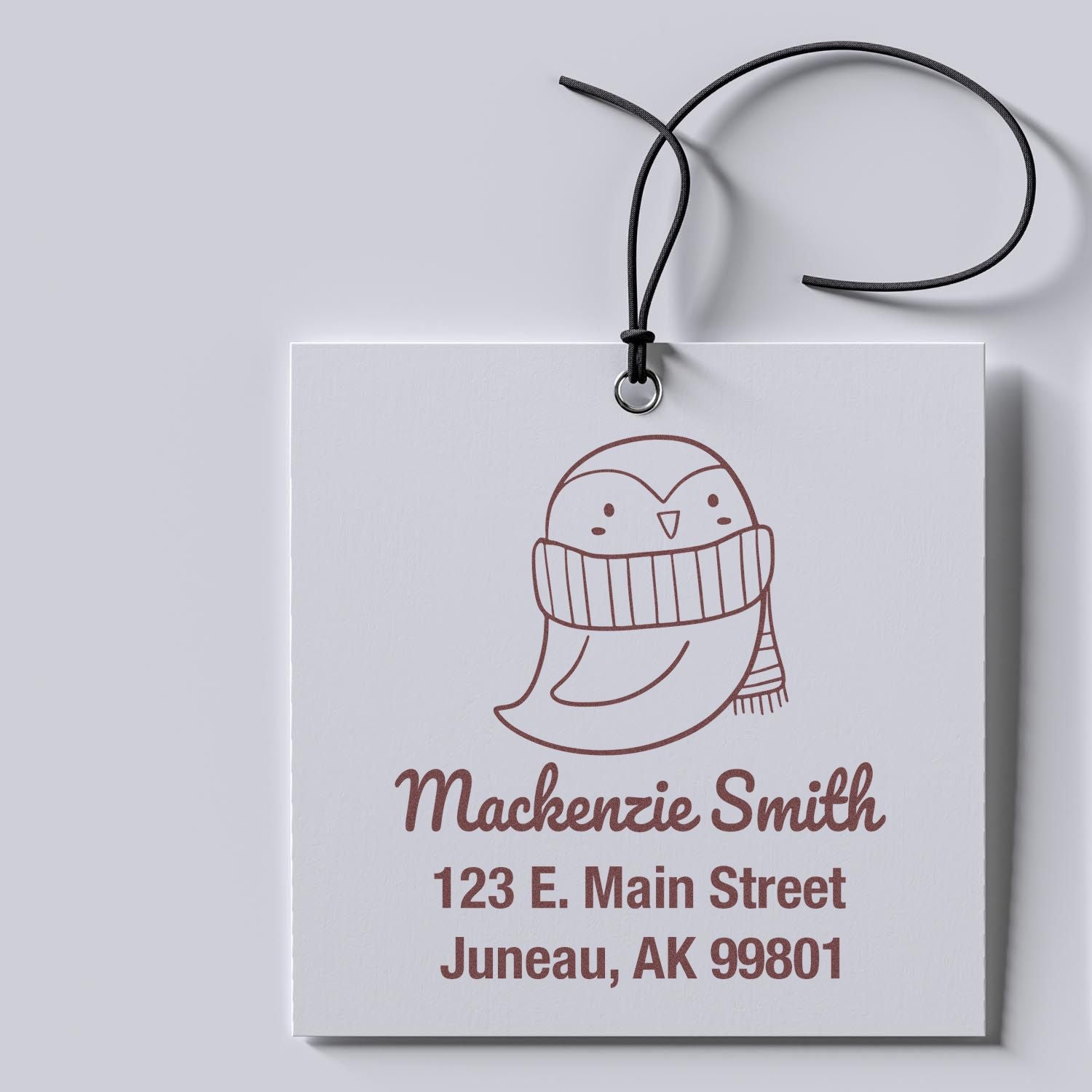 Portable Pre-Inked Pretty Penguin Custom Designed Return Address Stamper in use, featuring a cute penguin illustration. The Customized Return Mail Stamp displays "Mackenzie Smith, 123 E. Main Street, Juneau, AK 99801" on a white tag with a black string.