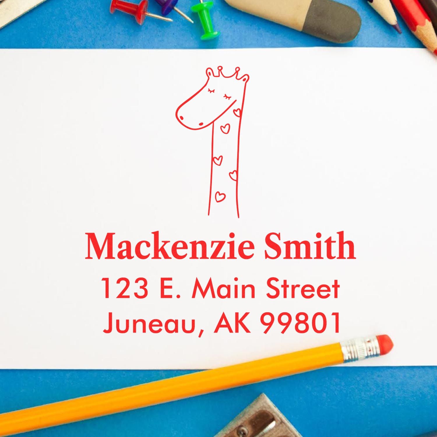 Portable Pre-Inked Lovely Giraffe Custom Designed Address Stamp on white paper with "Mackenzie Smith, 123 E. Main Street, Juneau, AK 99801" in red. Personalizable Mailing Stamper surrounded by colorful stationery on a blue background.