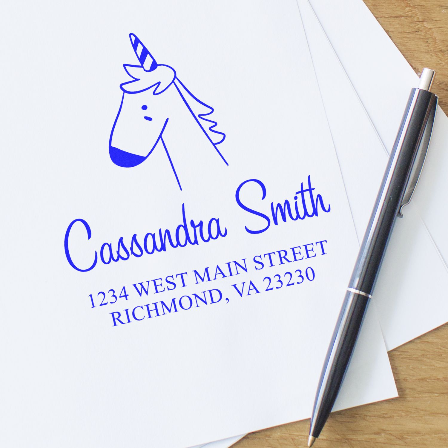 Self-Inking Cartoon Animal Whimsical Unicorn Custom Made Mailing Stamper on white paper with blue ink. The Customizable Home Stamp features a unicorn design, personalized with a name and address, next to a pen on a wooden surface.