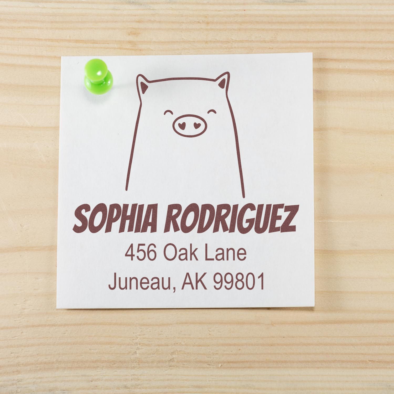 Image of a Portable Pre-Inked Cheery Pig Custom Designed Mailing Stamp on a wooden surface. The stamp features a cute pig illustration above the name "Sophia Rodriguez" and an address. This Custom Mailing Stamp is pinned with a green pushpin.