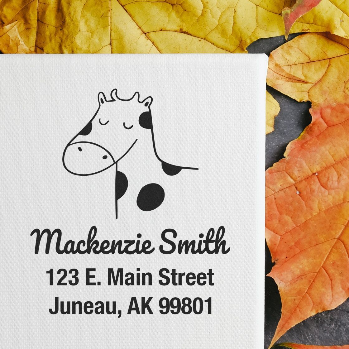 Wood handle stamp featuring a cartoon cow with the text "Mackenzie Smith, 123 E. Main Street, Juneau, AK 99801" on a canvas. Surrounded by autumn leaves. Keywords: Wood Handle Cow Cuddles Cartoon Animal Made To Order Home Stamper, Personalizable Home Stamp.