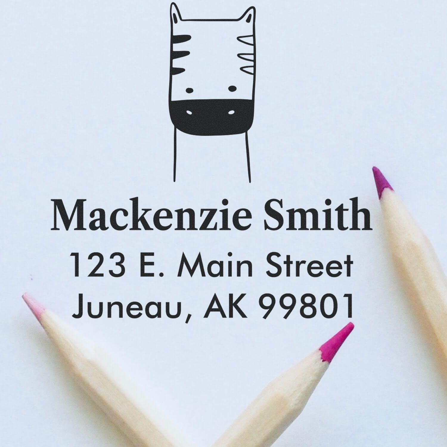Wood handle frolicsome zebra cartoon animal made to order envelope stamp with custom new address stamp for Mackenzie Smith, 123 E. Main Street, Juneau, AK 99801, shown with two pencils on a light background.