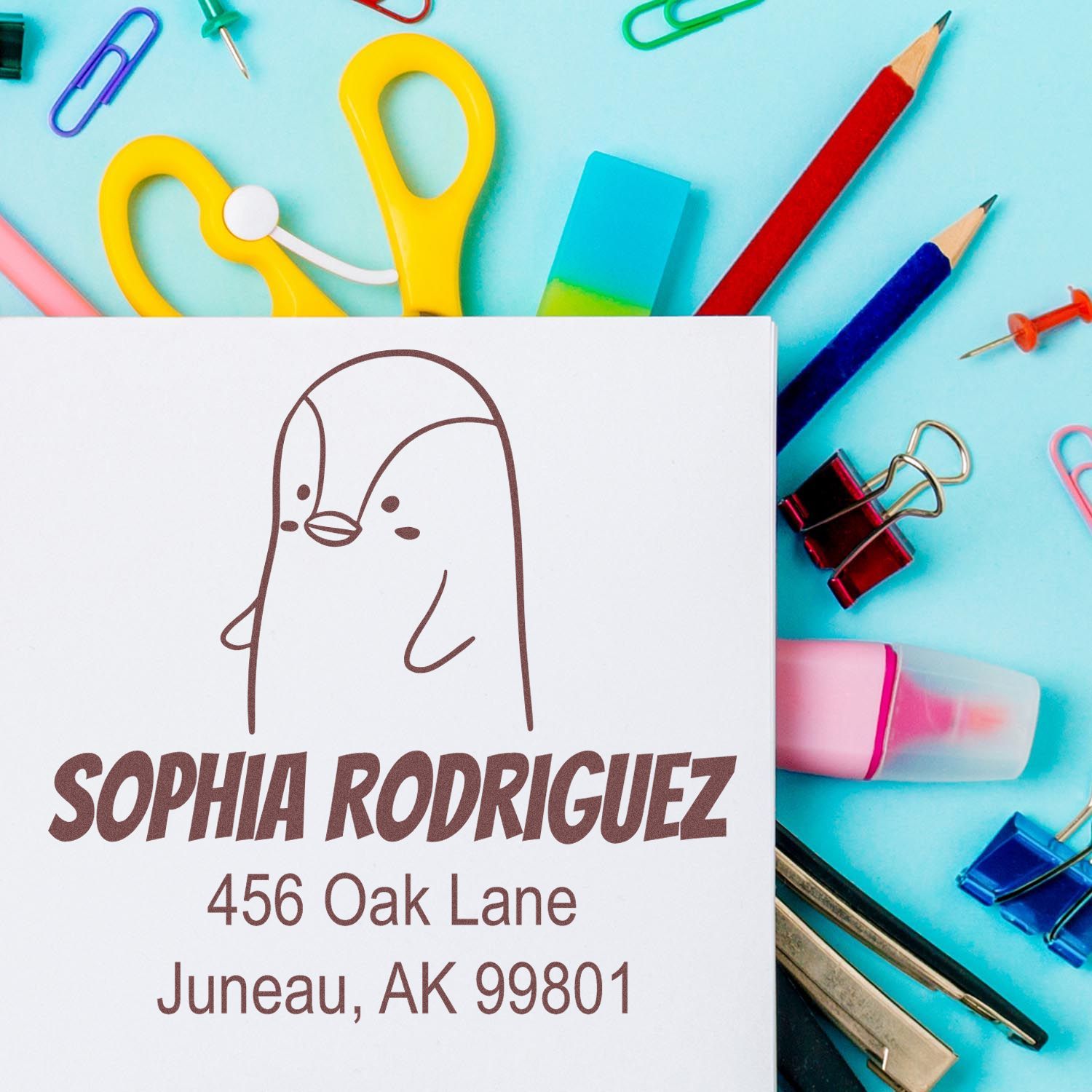 Self-Inking Cartoon Animal Perky Penguin Custom Made Envelope Stamper on a white envelope with the name and address "Sophia Rodriguez, 456 Oak Lane, Juneau, AK 99801." Surrounded by colorful stationery. Customized Mailing Stamp in use.