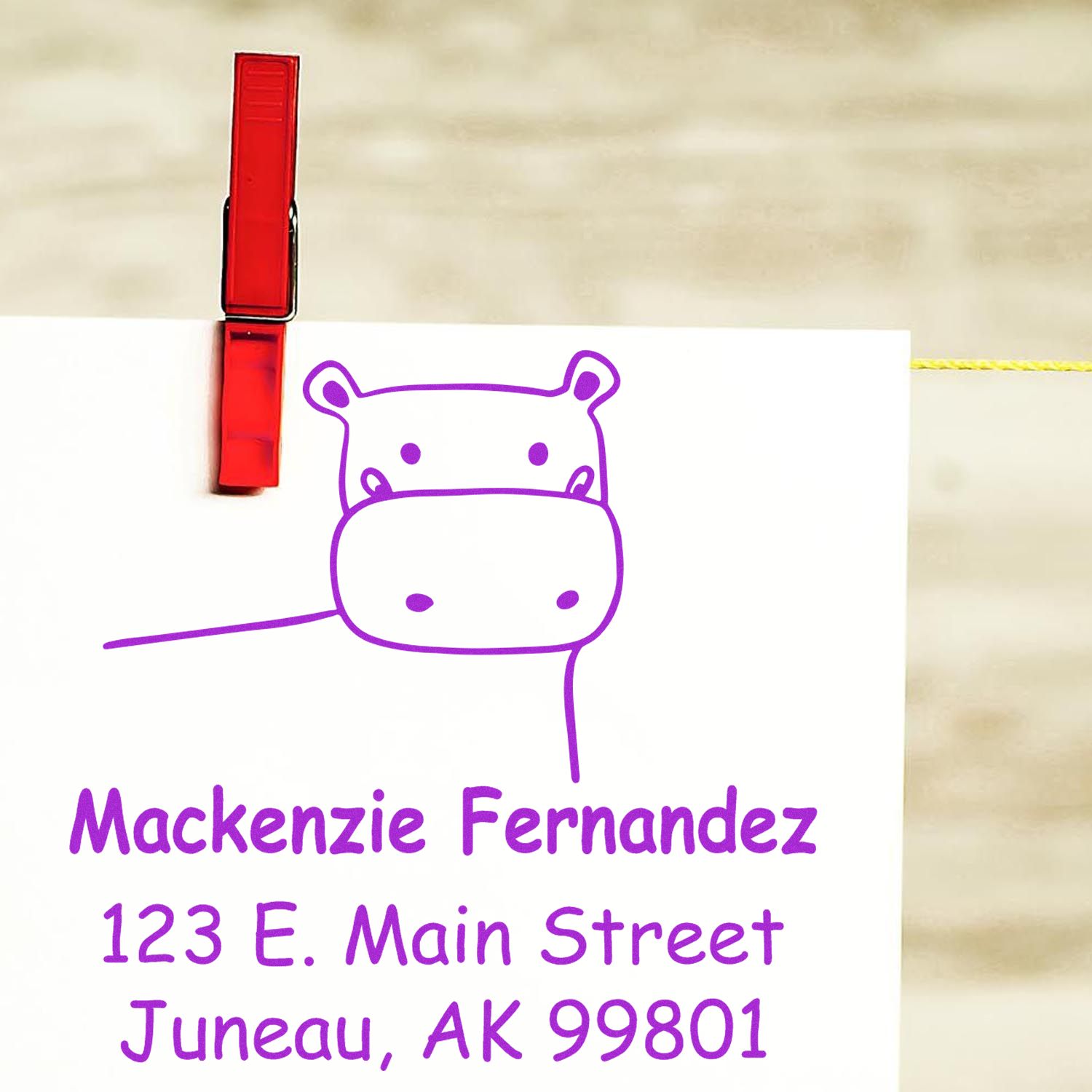 Wood handle happy hippo cartoon animal made to order address stamper on a personalized return mail stamp. The stamp features a cute hippo design with purple text displaying a name and address, clipped to a line with a red clothespin.