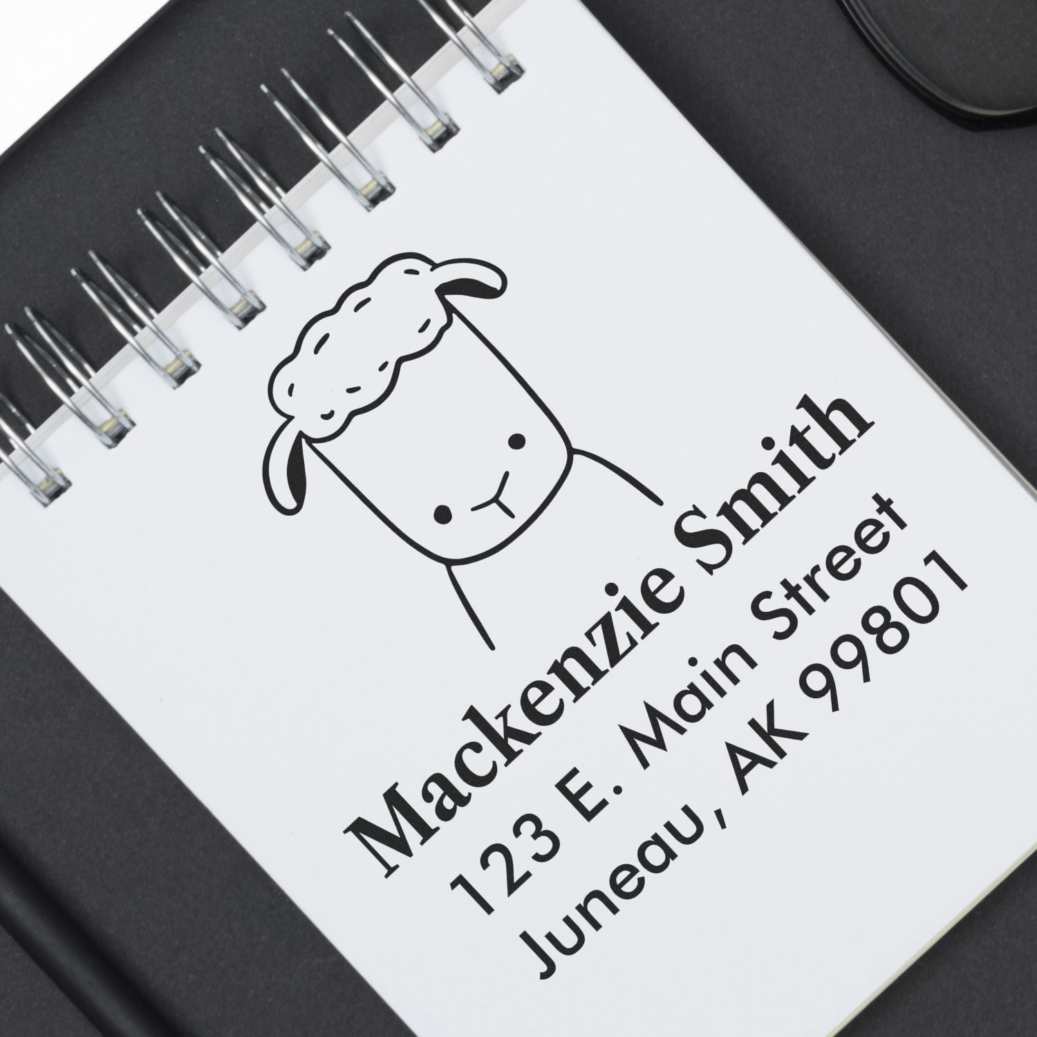 Wood handle fluffy sheep cartoon animal made to order name and address stamper used on a notebook. The custom home stamp displays "Mackenzie Smith, 123 E. Main Street, Juneau, AK 99801" with a cute sheep illustration above the text.