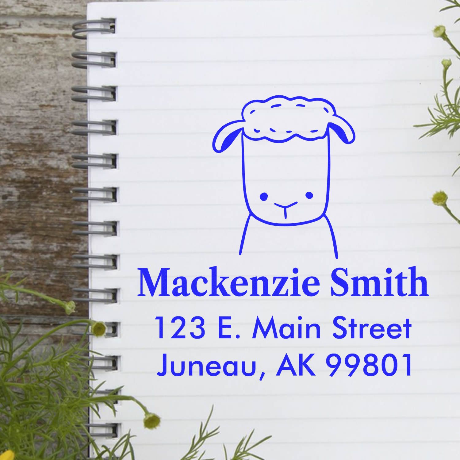 A notebook page with a blue Fluffy Sheep Pre-Inked Customized Address Stamper design, featuring a sheep illustration above the name "Mackenzie Smith" and address "123 E. Main Street, Juneau, AK 99801." Custom-Made Home Stamper in use.