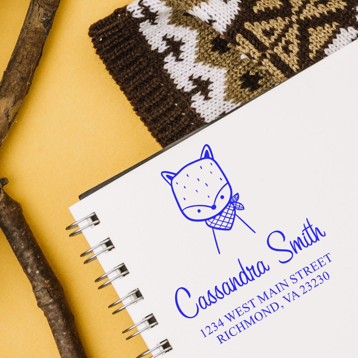 Self-inking cartoon animal fancy fox custom made name and address stamper on a notebook. The customized home stamp displays "Cassandra Smith, 1234 West Main Street, Richmond, VA 23230" in blue ink, next to a knitted item and twigs.