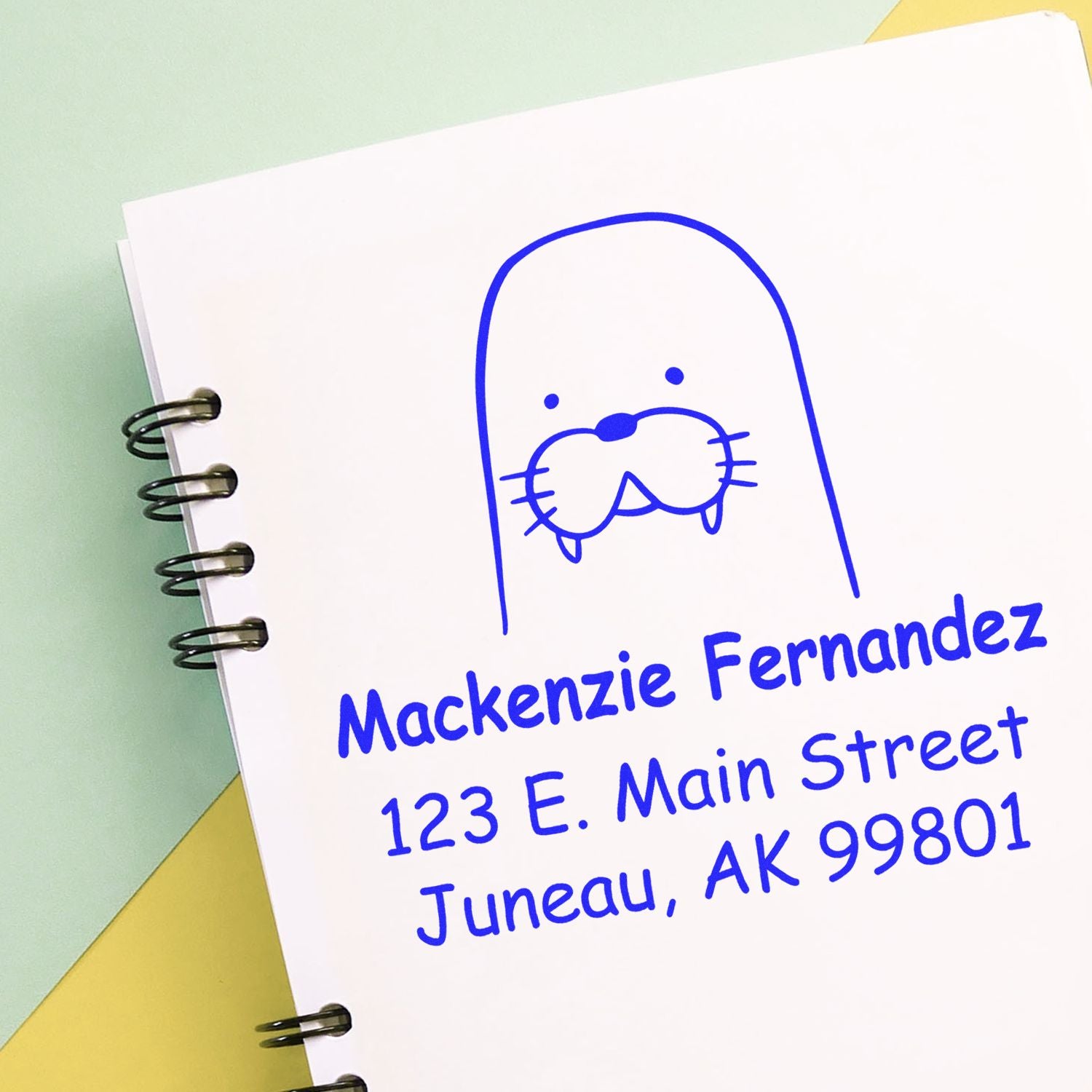 A Portable Pre-Inked Wacky Walrus Custom Designed Name And Address Stamper is shown on a notebook. The stamp displays "Mackenzie Fernandez, 123 E. Main Street, Juneau, AK 99801" in blue ink. This Customizable Return Mail Stamp features a walrus graphic.