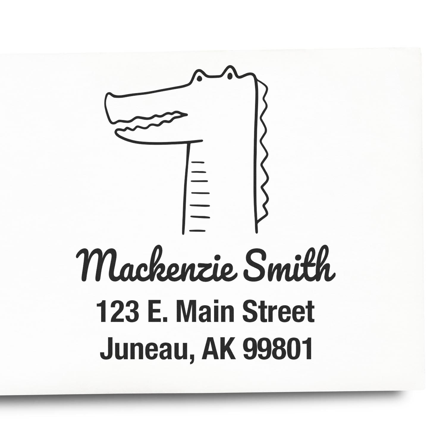 A customized mailing stamp featuring a bright-eyed crocodile cartoon with a wood handle. The stamp displays the name Mackenzie Smith and address 123 E. Main Street, Juneau, AK 99801. It's a made-to-order home address stamper.