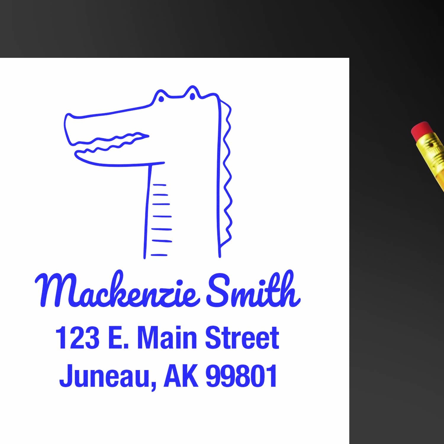 Wood handle bright-eyed crocodile cartoon animal made to order home address stamper on white paper. Customized return mail stamp with blue text reads "Mackenzie Smith, 123 E. Main Street, Juneau, AK 99801." Pencil with eraser visible on the side.