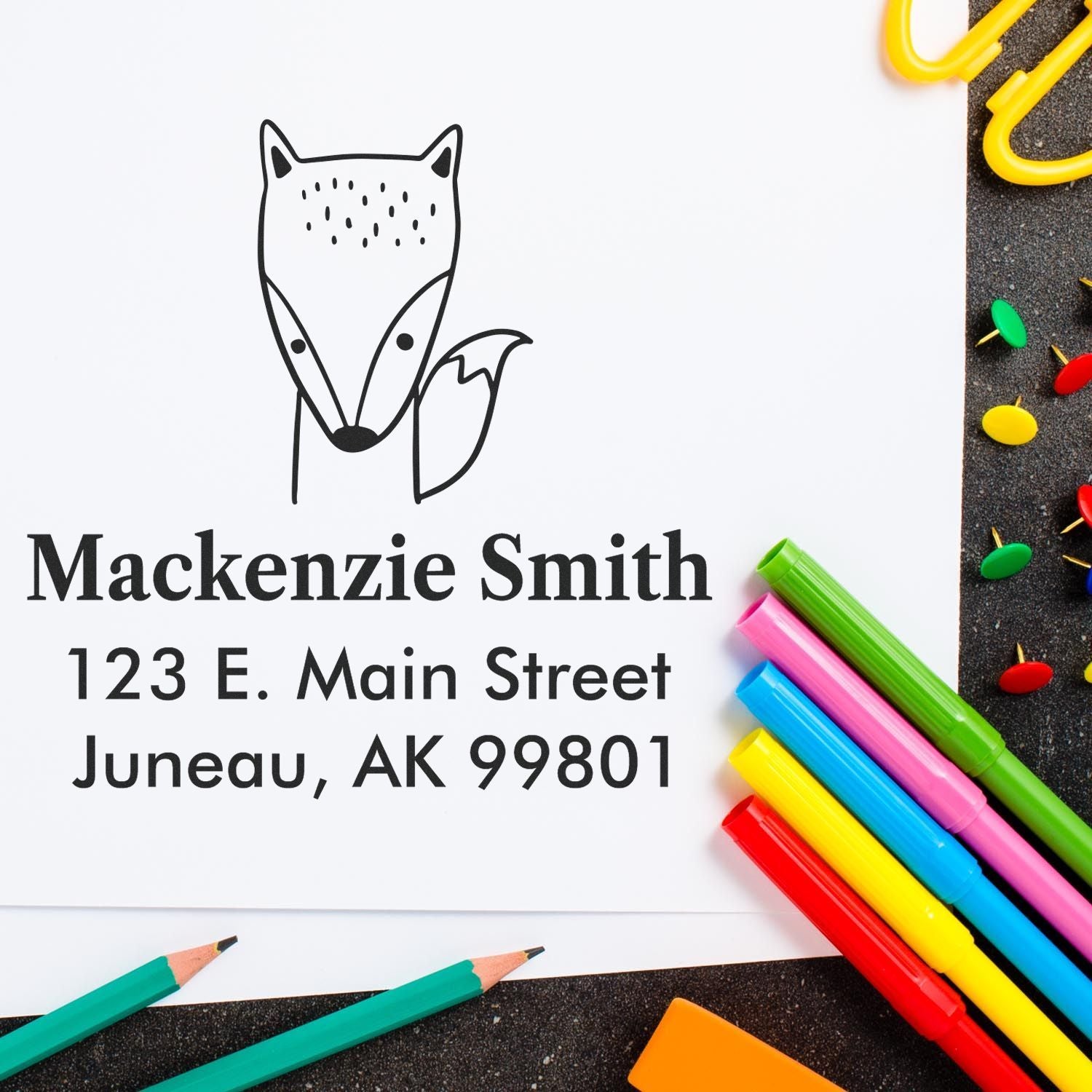 Wood handle fast fox cartoon animal made to order return address stamp on paper with text "Mackenzie Smith, 123 E. Main Street, Juneau, AK 99801." Custom new address stamp shown with colorful pens and paperclips on a dark surface.
