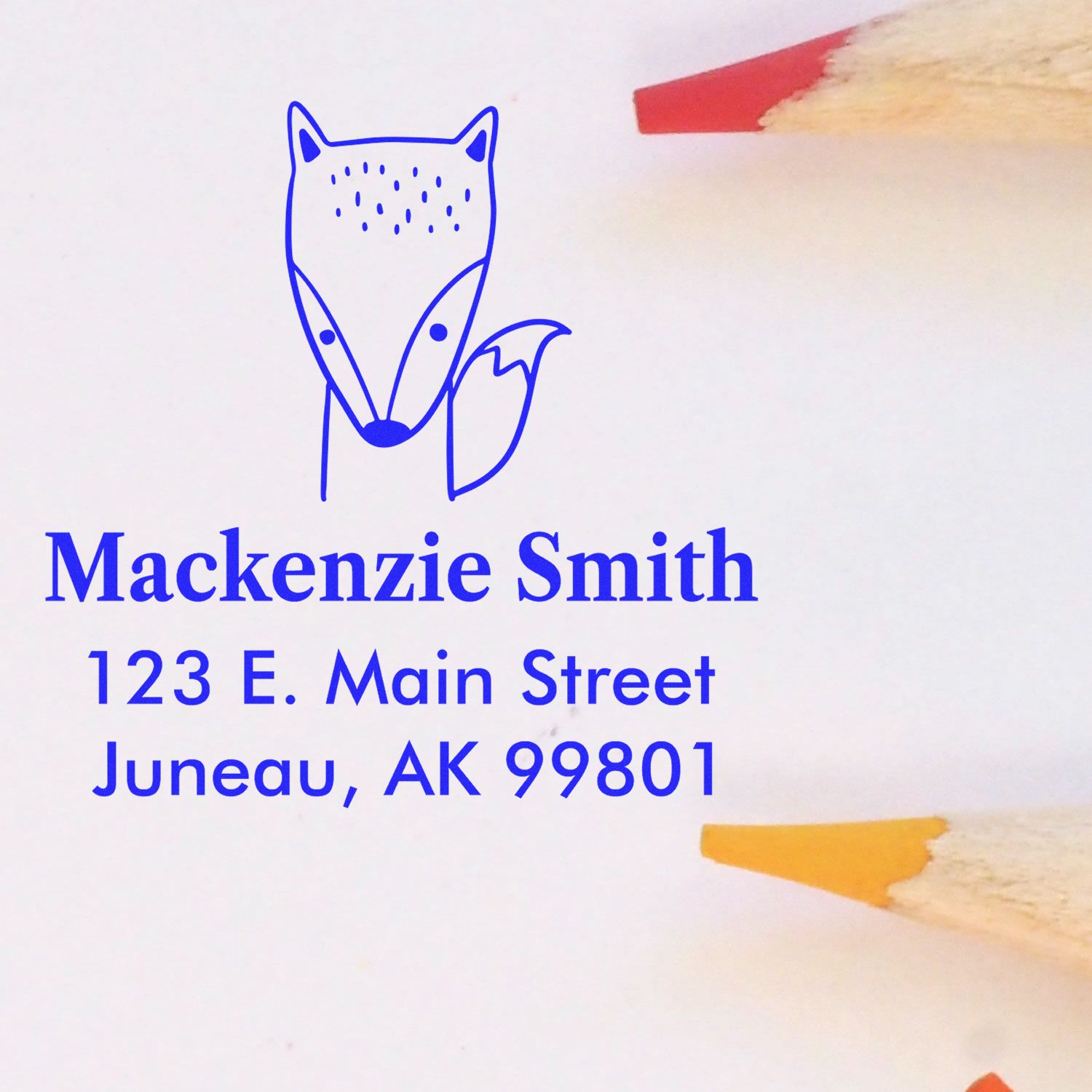 Self-inking cartoon animal fast fox custom-made home address stamper featuring a blue fox design. Text reads "Mackenzie Smith, 123 E. Main Street, Juneau, AK 99801." Custom-made return mail stamp shown with colored pencils nearby.