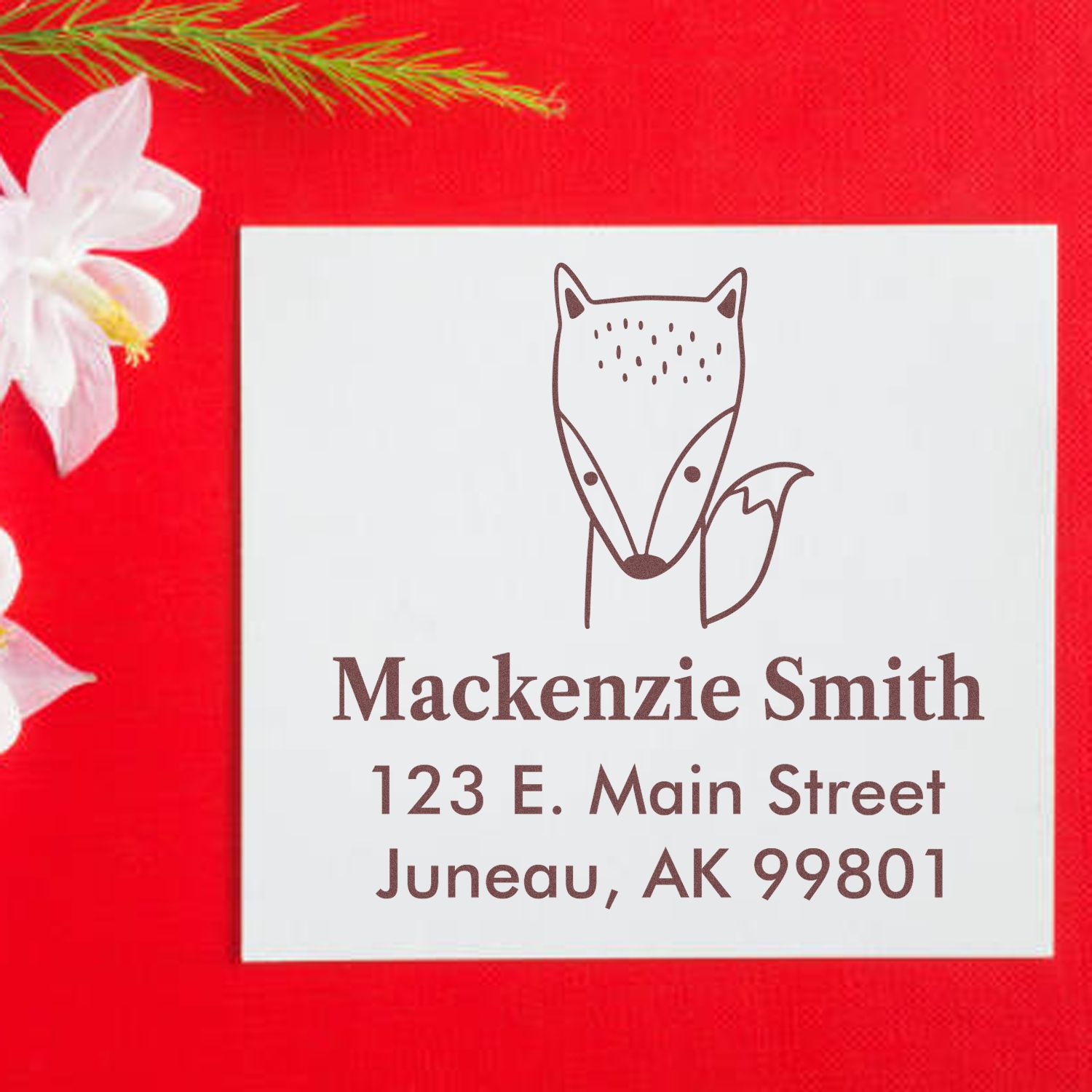Fast Fox Pre-Inked Customized Home Address Stamp on a white envelope with a fox design, labeled "Mackenzie Smith, 123 E. Main Street, Juneau, AK 99801." The Custom Return Mail Stamper is set against a red background with white flowers.