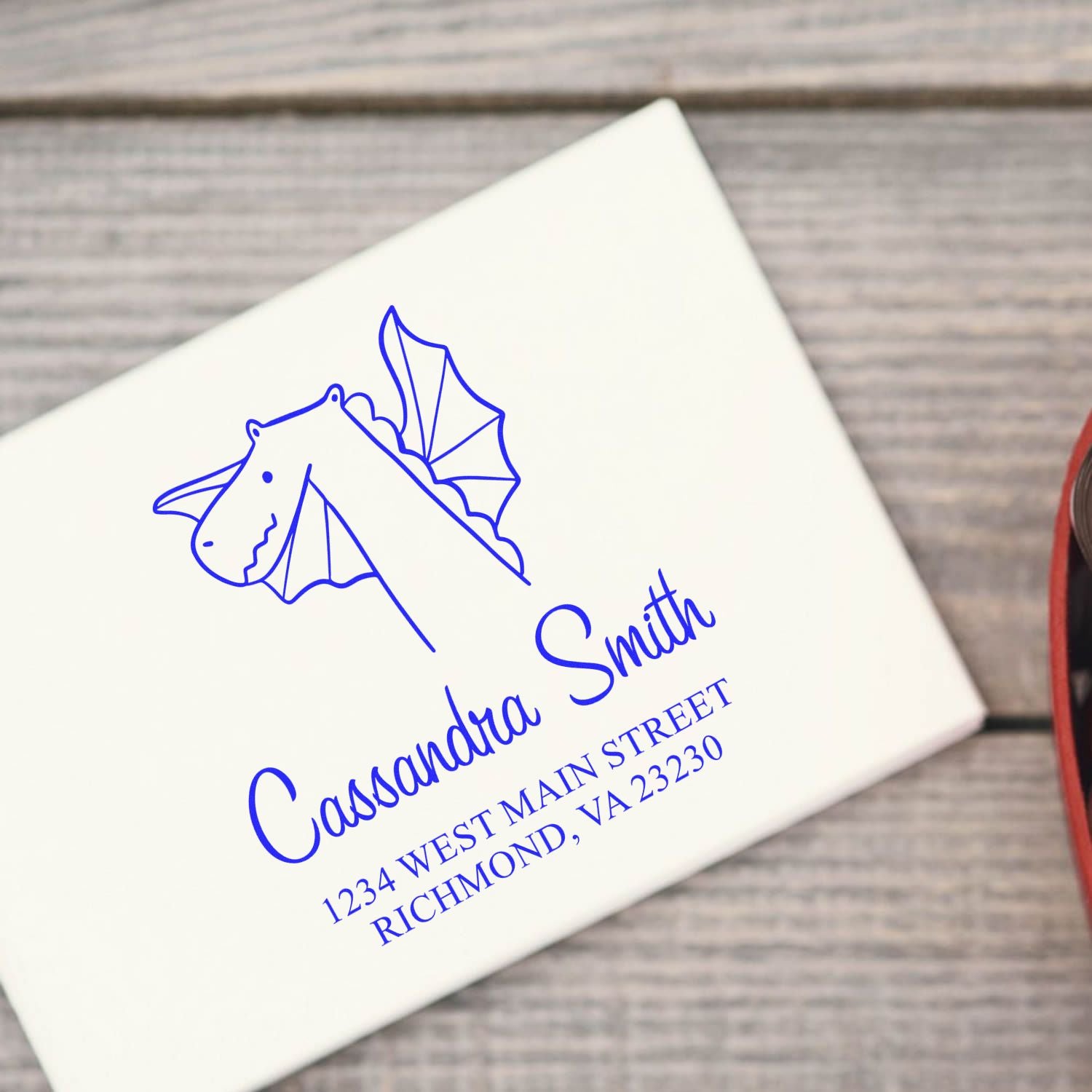 Wood handle dragon delight cartoon animal made to order return address stamper on a white envelope. The design features a dragon with wings, and the text is personalizable for a new address stamp. The envelope is placed on a wooden surface.