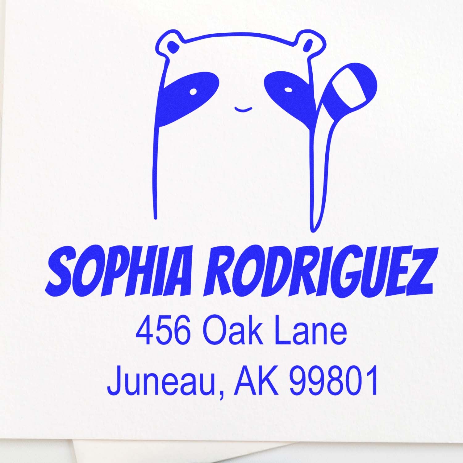 Renegade Racoon Pre-Inked Customized Return Address Stamp featuring a cute raccoon design with the name "Sophia Rodriguez" and address "456 Oak Lane, Juneau, AK 99801" in blue. Ideal as a custom mailing stamp for personalized correspondence.