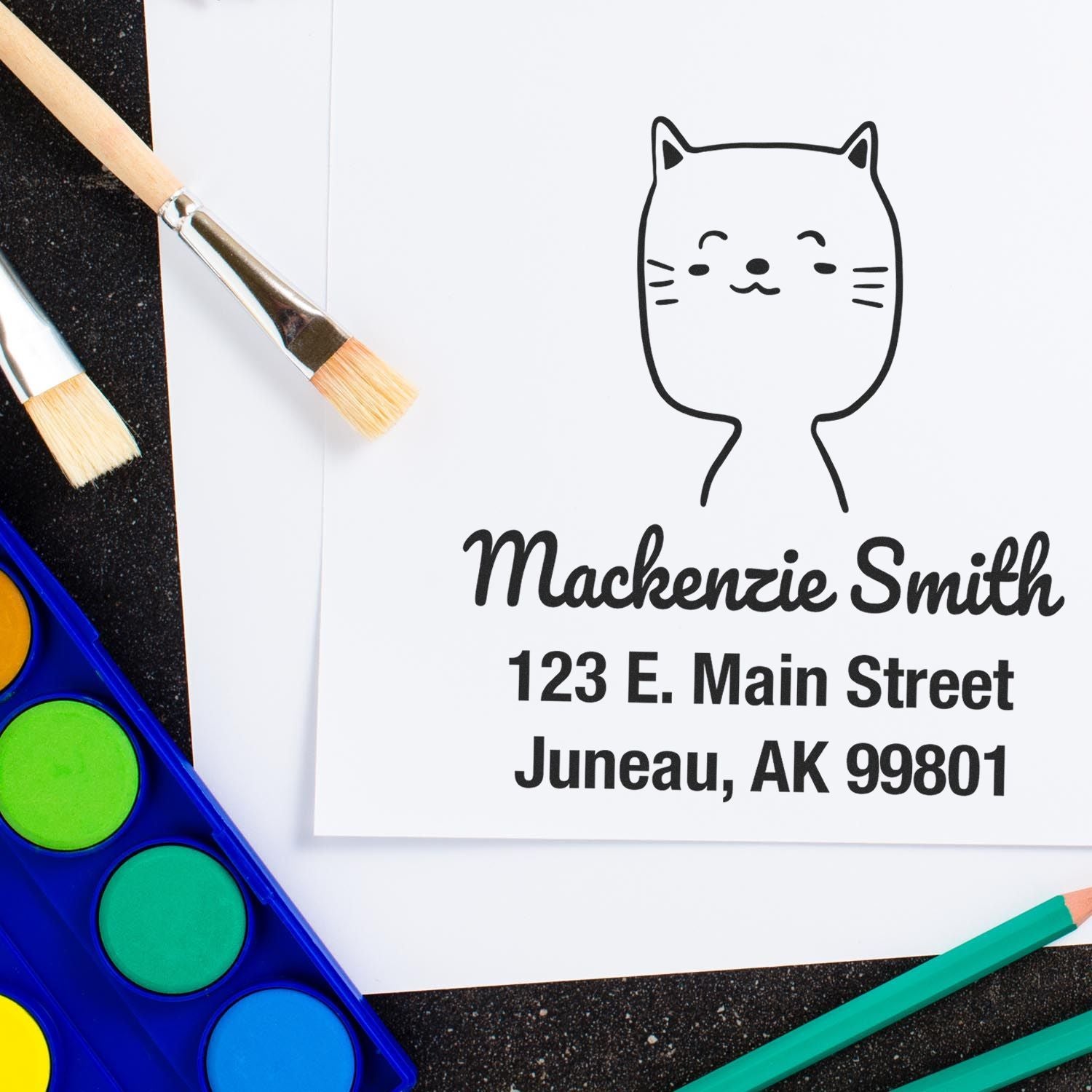 Wood handle charming cat cartoon animal custom mail stamp on paper with "Mackenzie Smith, 123 E. Main Street, Juneau, AK 99801" text. Custom mailing stamp design with paintbrushes and watercolor palette nearby.