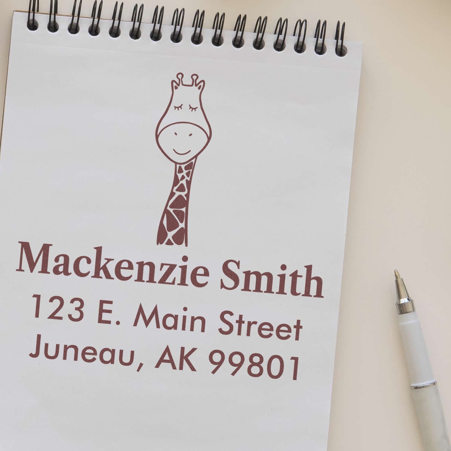 Wood Handle Jolly Giraffe Cartoon Animal Custom Mail Stamper imprint on a notepad, featuring a giraffe design above the name "Mackenzie Smith" and address "123 E. Main Street, Juneau, AK 99801." Custom-Made Address Stamp next to a pen.