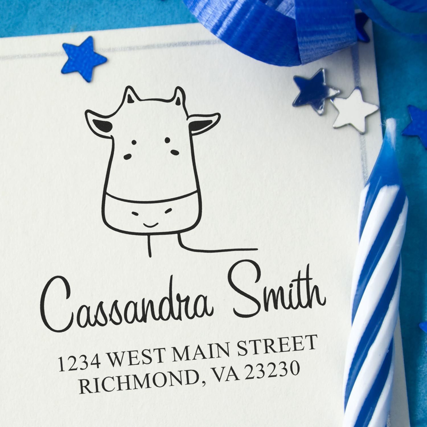 Wood Handle Giraffe Grins Cartoon Animal Custom Return Address Stamp on a white envelope with "Cassandra Smith, 1234 West Main Street, Richmond, VA 23230." Blue ribbon and candle nearby. Customizable New Address Stamp for personalized mail.