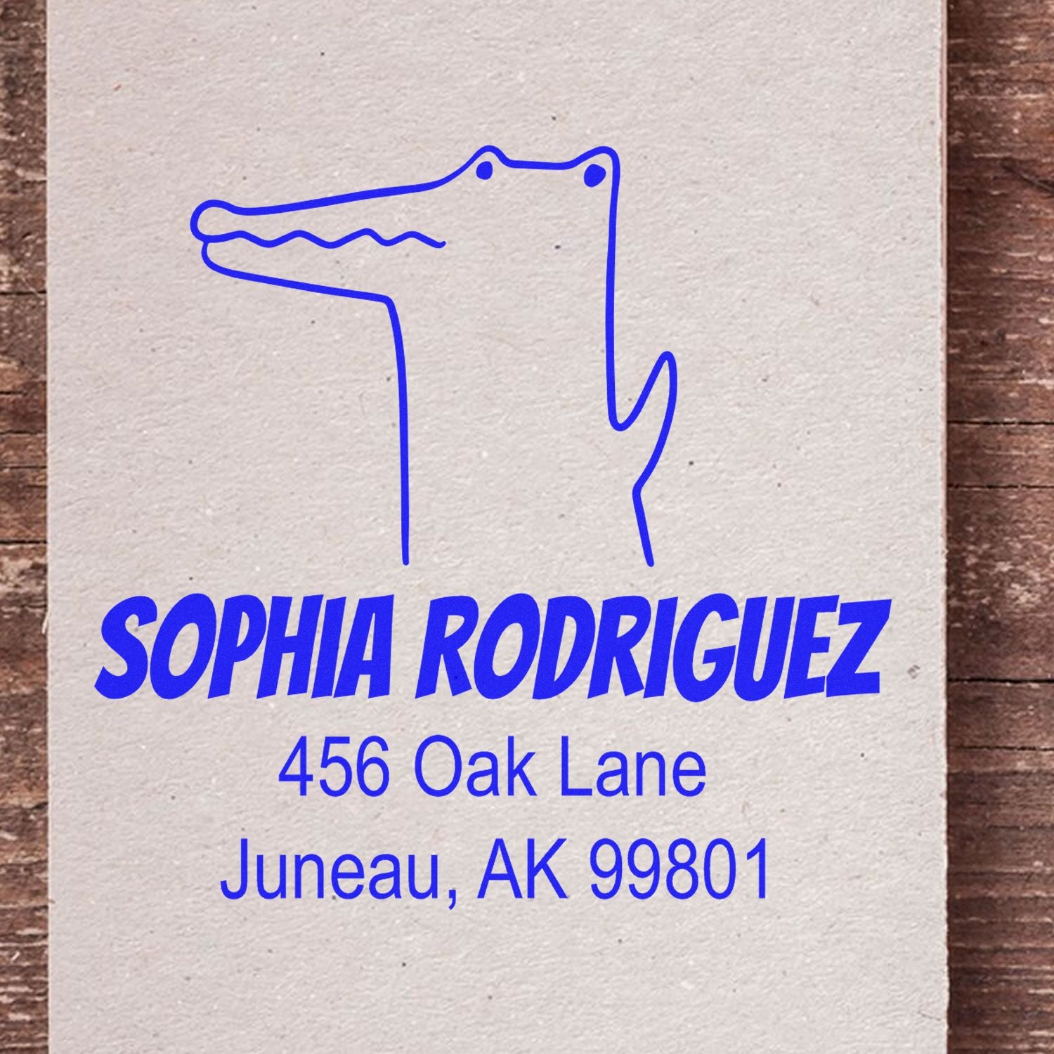 Portable Pre-Inked Charming Crocodile Made To Order Mail Stamp featuring a blue crocodile design above the name Sophia Rodriguez and address 456 Oak Lane, Juneau, AK 99801. Custom Return Mail Stamper on a wooden background.