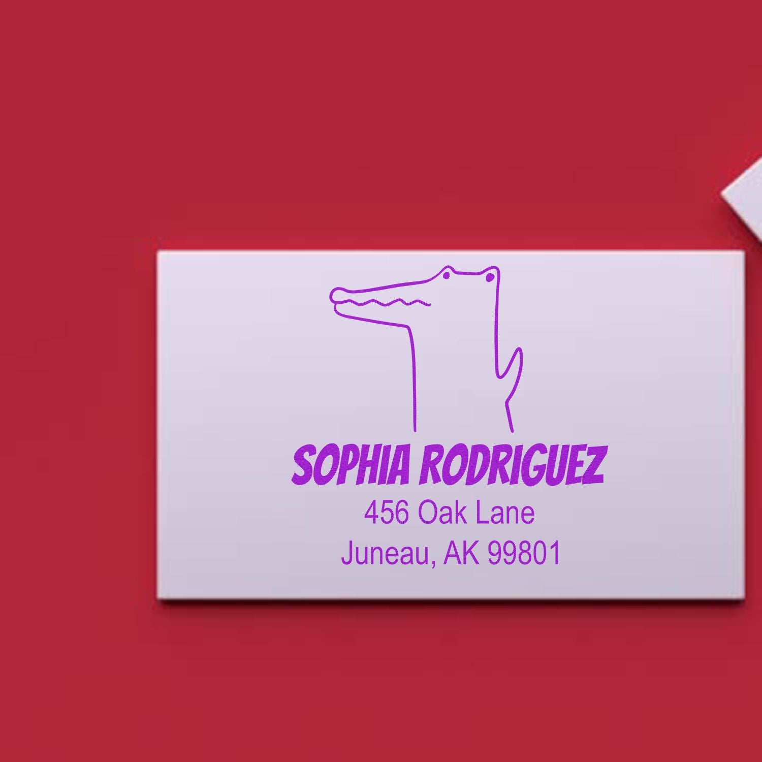 Image of a white envelope on a red background featuring a purple Portable Pre-Inked Charming Crocodile Made To Order Mail Stamp. The stamp includes a custom home stamper design with the name "Sophia Rodriguez" and an address in Juneau, AK.
