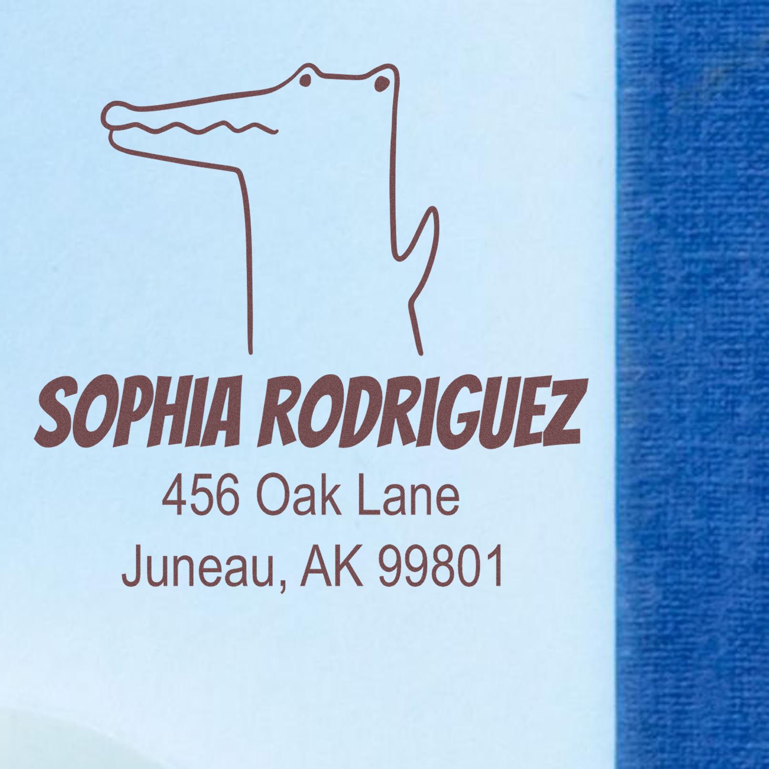 Wood handle charming crocodile cartoon animal custom return address stamper on blue paper. Personalized new address stamp with crocodile illustration and text: "Sophia Rodriguez, 456 Oak Lane, Juneau, AK 99801."