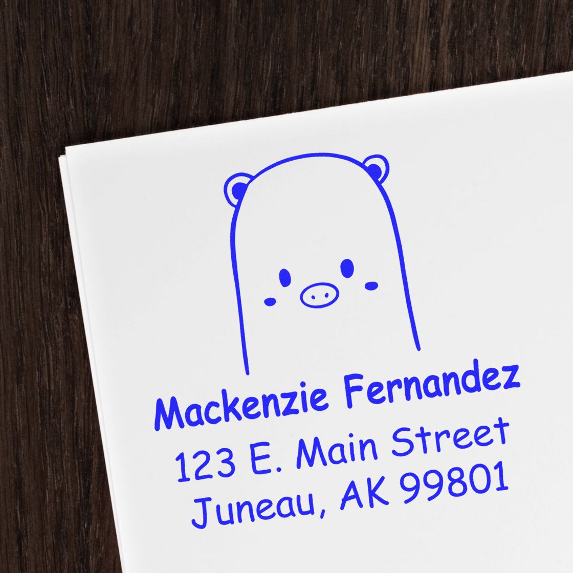 Image of a Portable Pre-Inked Beautiful Bear Made To Order Mail Stamper on a white envelope. The Customized Home Stamp features a cute bear design with the name "Mackenzie Fernandez" and address in blue ink, placed on a dark wooden surface.