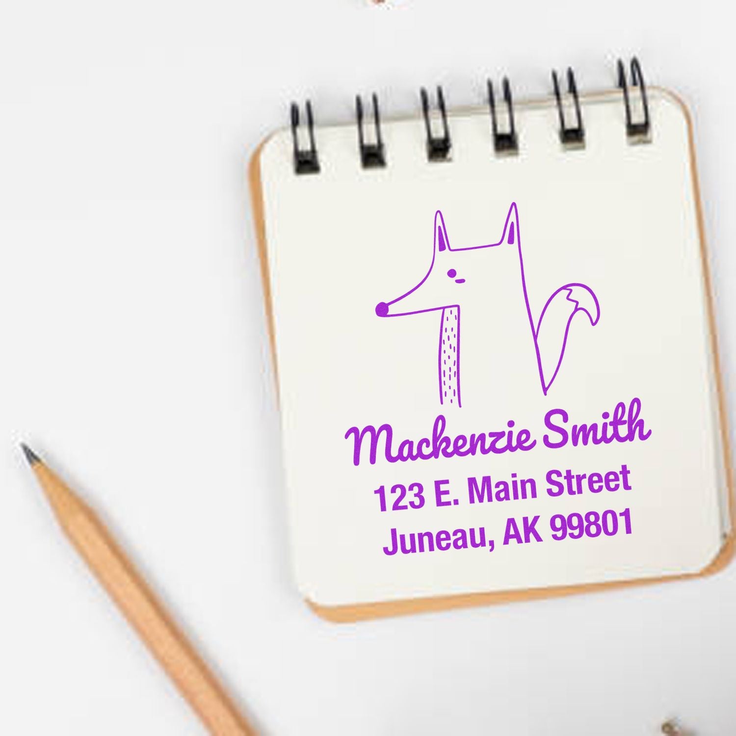 Wood Handle Finicky Fox Cartoon Animal Custom Address Stamper used on a notepad, displaying a purple fox illustration with the text "Mackenzie Smith, 123 E. Main Street, Juneau, AK 99801." Customized Home Stamp next to a wooden pencil.