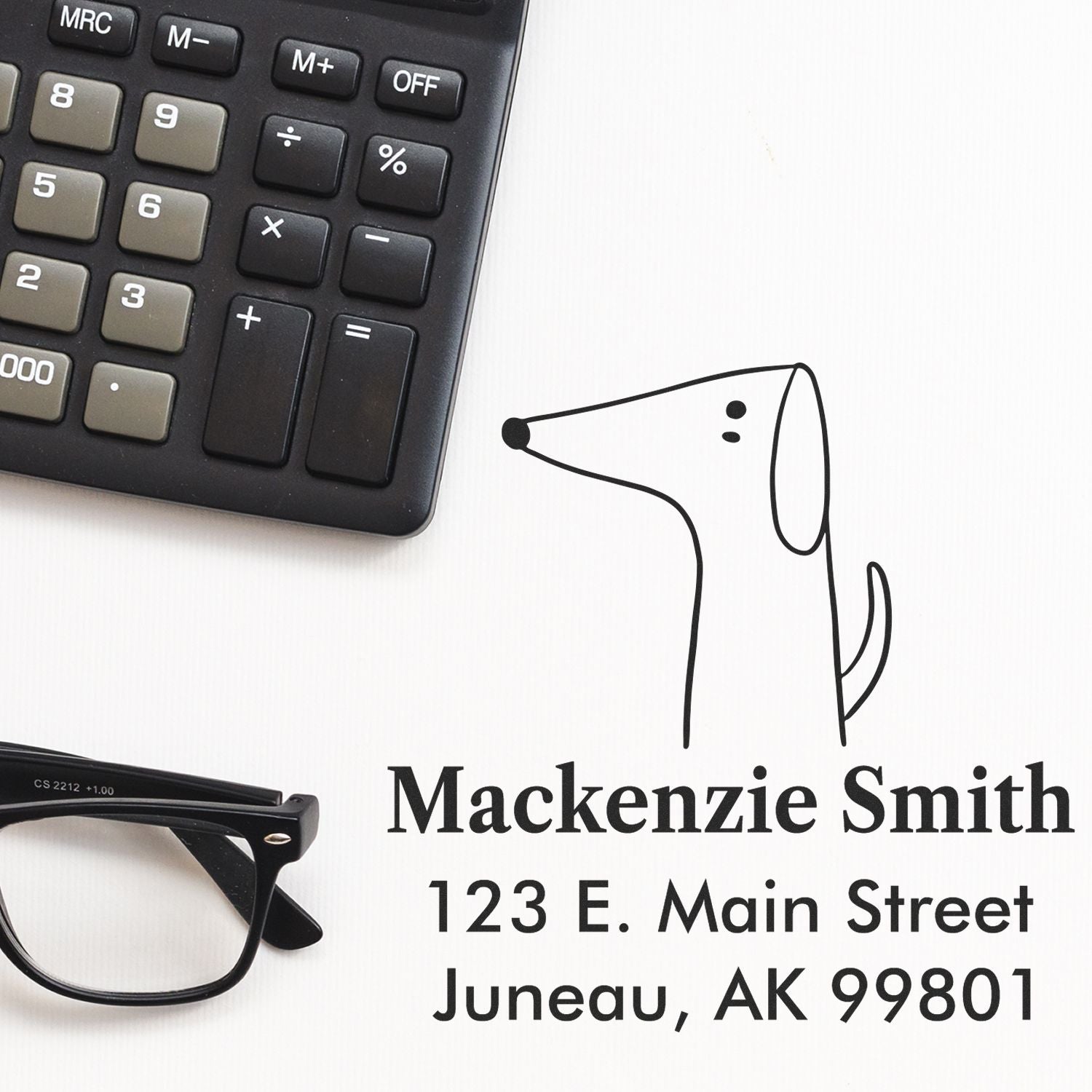 Image of a Portable Pre-Inked Perky Pooch Made To Order Return Address Stamper in use. The Personalizable Address Stamp displays "Mackenzie Smith, 123 E. Main Street, Juneau, AK 99801" alongside a simple dog illustration, next to a calculator and glasses.