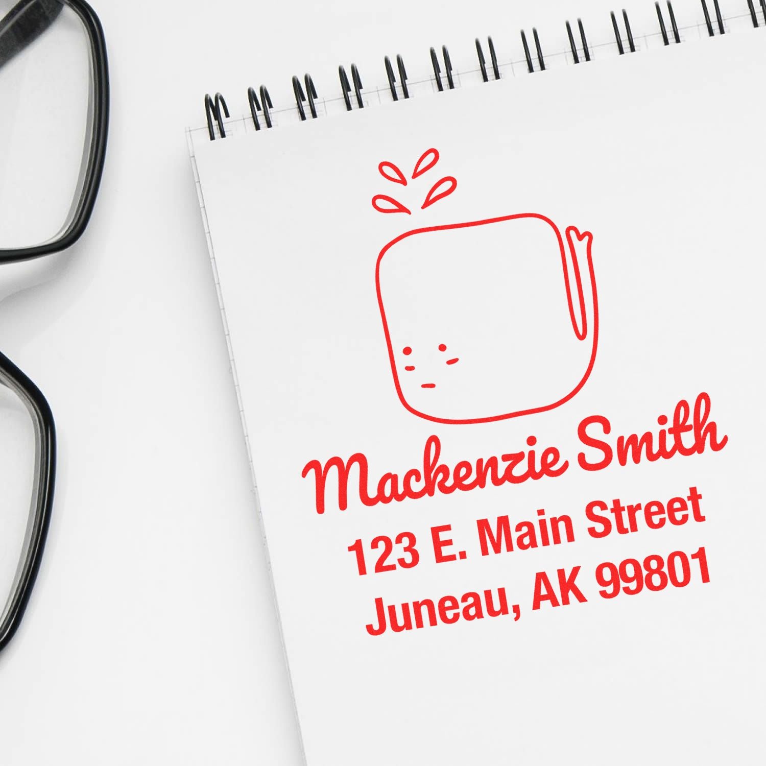 Joyful Whale Pre-Inked Handcrafted Address Stamp in use on a notepad, displaying a customizable mailing stamp with a red whale design and the address "Mackenzie Smith, 123 E. Main Street, Juneau, AK 99801." Glasses are placed beside the notepad.