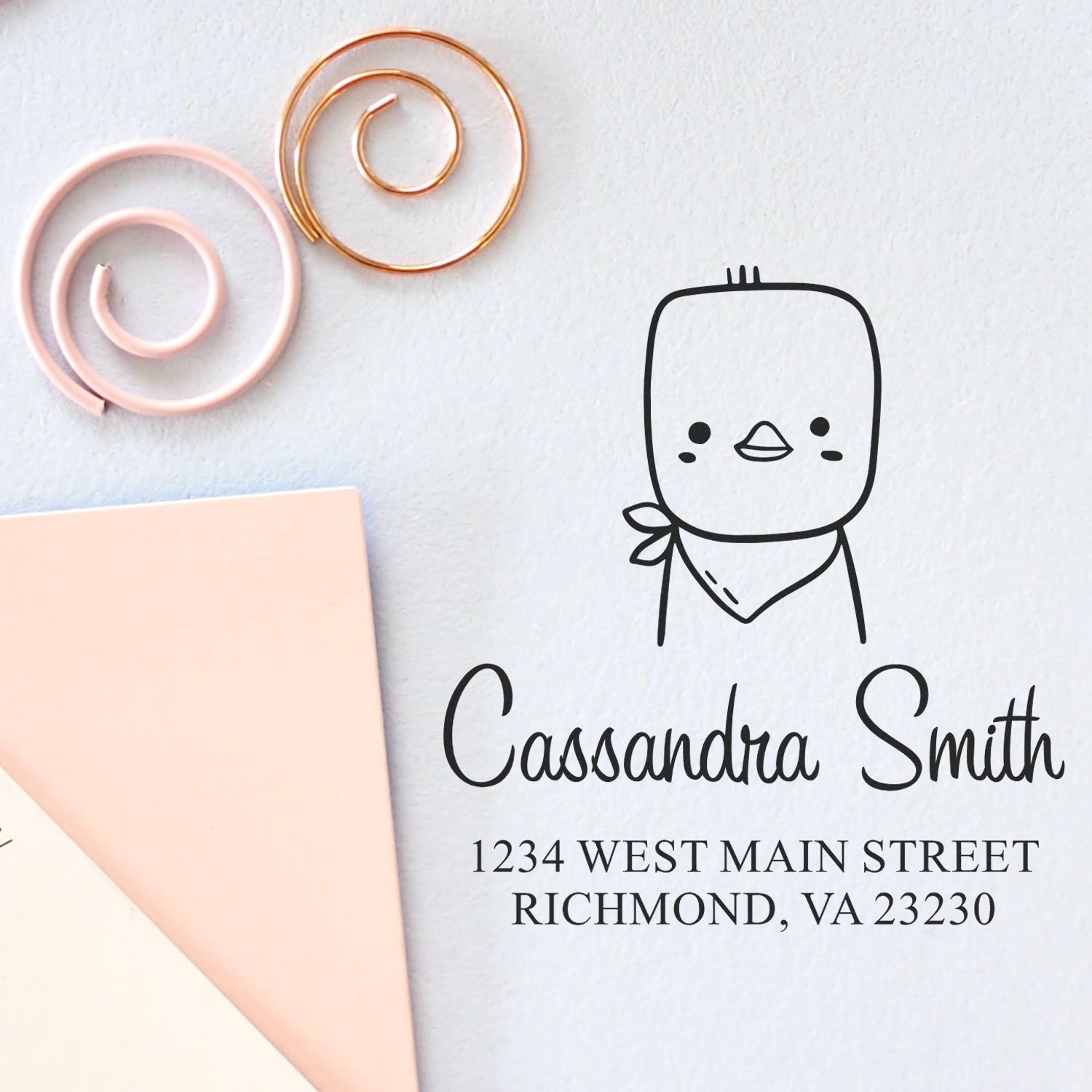 Wood handle stamp with a cute chick cartoon animal design on a custom envelope. The stamp features a custom home address: "Cassandra Smith, 1234 West Main Street, Richmond, VA 23230." Spiral paperclips are nearby.