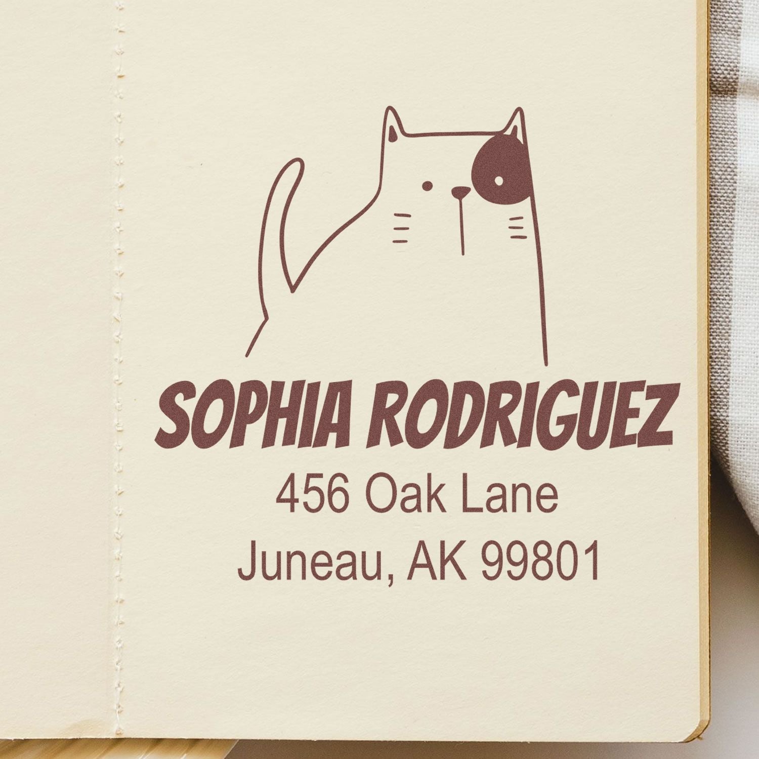Wood handle fluffy feline cartoon animal custom envelope stamper in use, displaying a customizable new address stamp with the name "Sophia Rodriguez" and address "456 Oak Lane, Juneau, AK 99801" on a beige paper background.