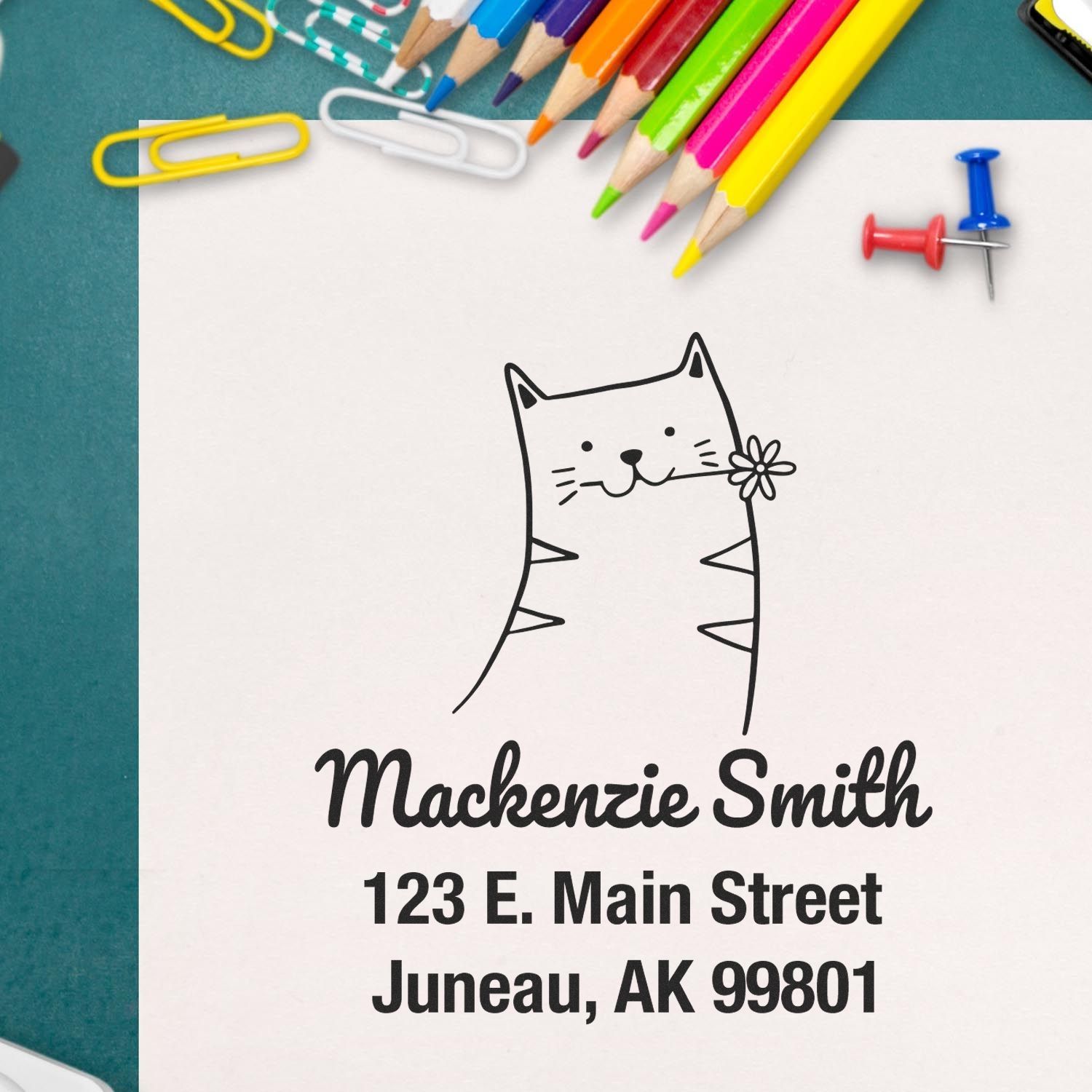 Wood Handle Happy Cat Cartoon Animal Custom Address Stamper on paper with "Mackenzie Smith, 123 E. Main Street, Juneau, AK 99801." Surrounded by colorful pencils and paperclips. Customizable Return Mail Stamp for personalized use.