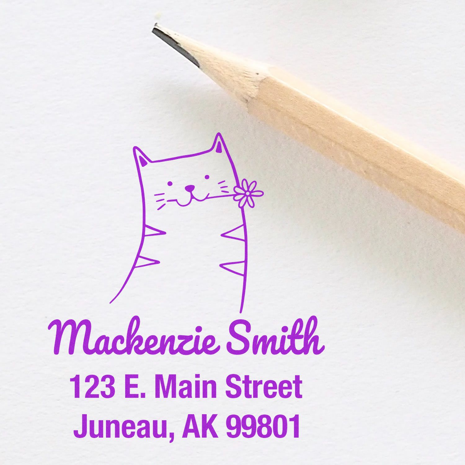 Portable Pre-Inked Happy Cat Made To Order Envelope Stamp with a cute cat design and flower, alongside a Custom New Address Stamper showing "Mackenzie Smith, 123 E. Main Street, Juneau, AK 99801" in purple ink, next to a wooden pencil.