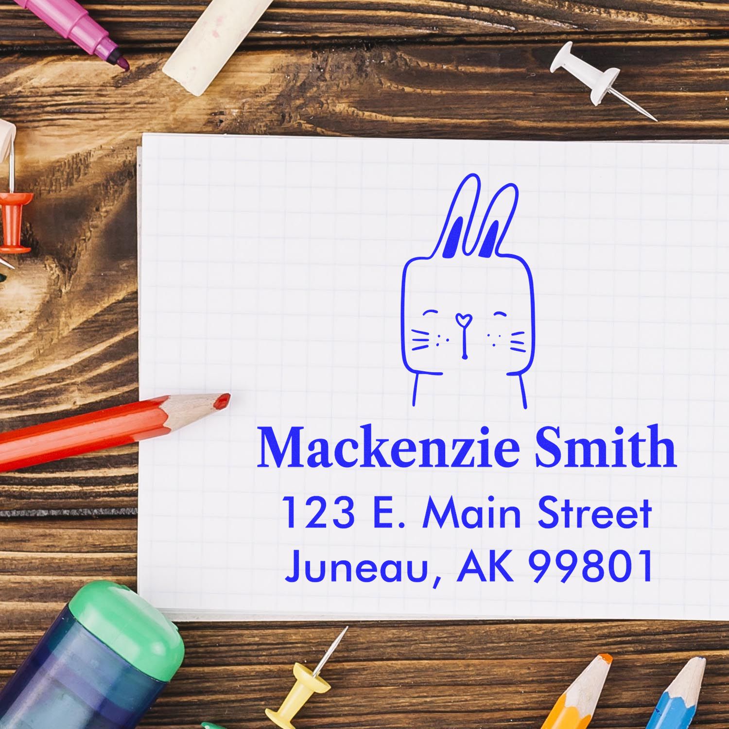 Wood Handle Fuzzy Rabbit Cartoon Animal Custom Name And Address Stamp on paper with "Mackenzie Smith, 123 E. Main Street, Juneau, AK 99801" in blue ink. Surrounded by colorful pencils and push pins on a wooden surface. Customizable Home Stamp.