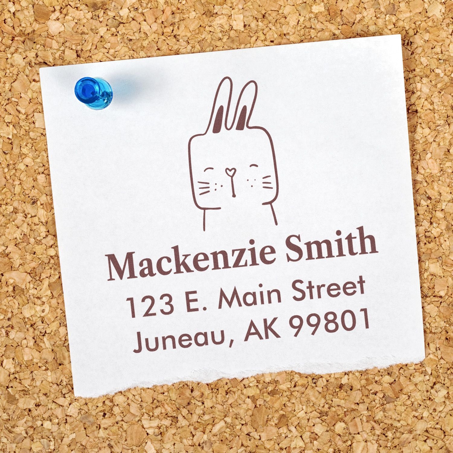 Fuzzy Rabbit Pre-Inked Handcrafted Address Stamp on a corkboard, featuring a cute rabbit design. The Customizable Return Mail Stamper displays the name "Mackenzie Smith" with an address in Juneau, AK. A blue pushpin secures the paper.